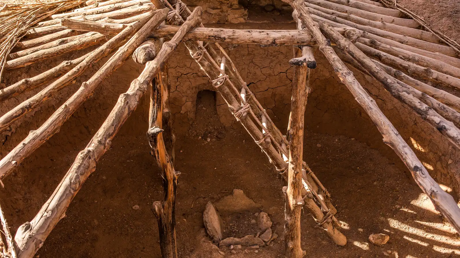 Wooden beams form the roof of a round, ancient pit house with an entry ladder inside.