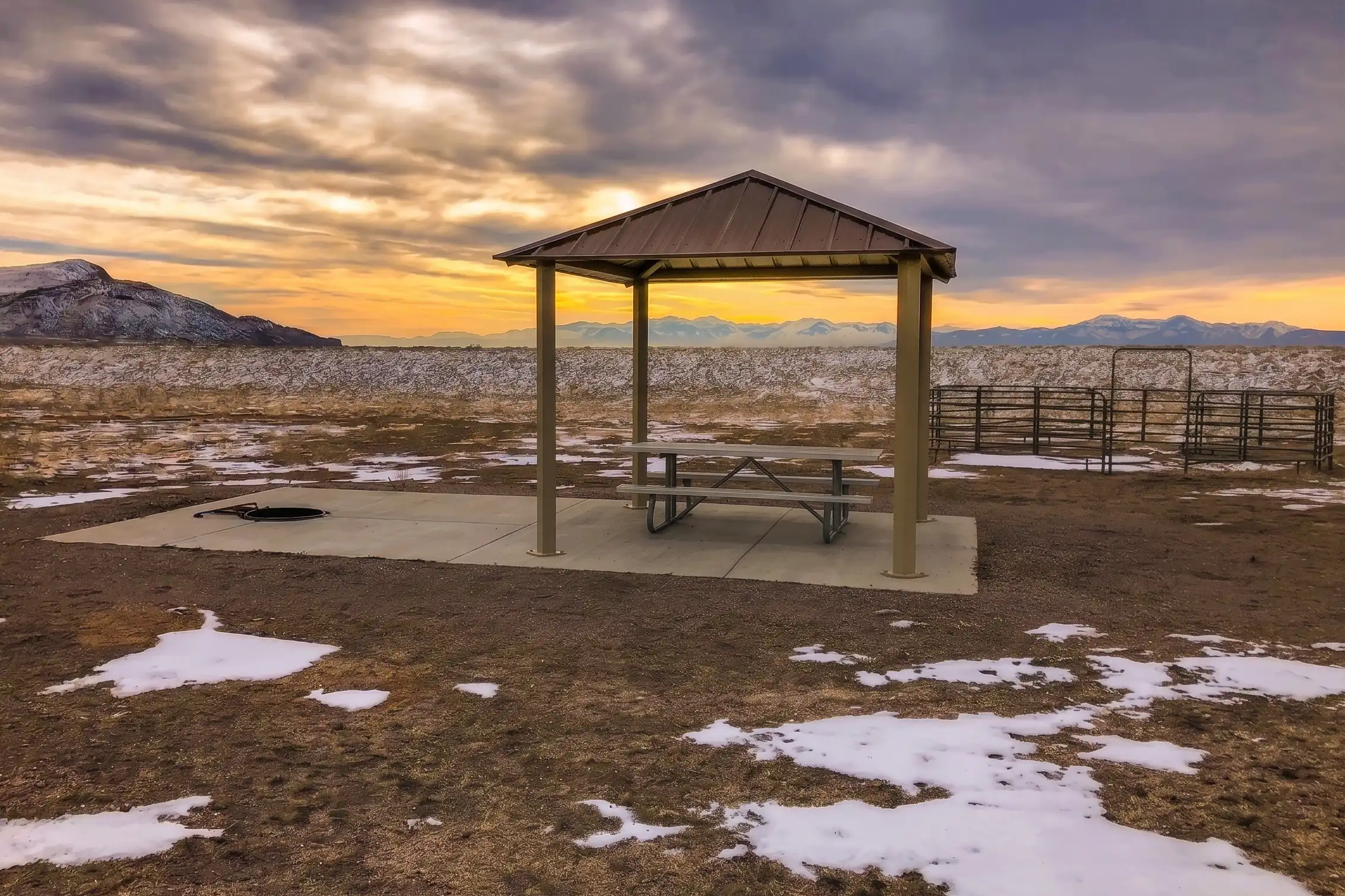 Picnic shelter on concrete slab in snowy field with mountains and sunset in background.