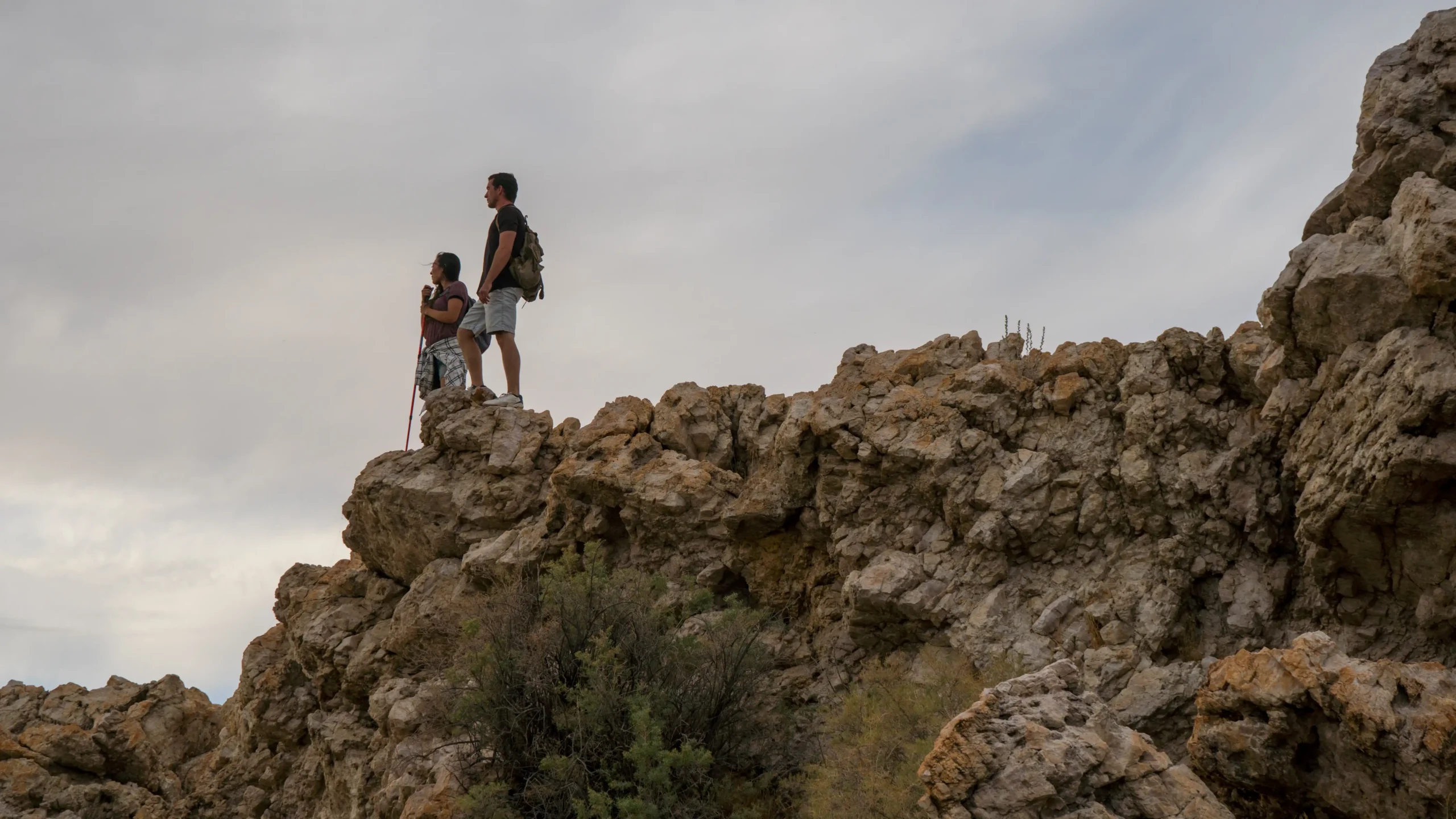 Two people stand on a rocky cliff, looking at the view under a cloudy sky.
