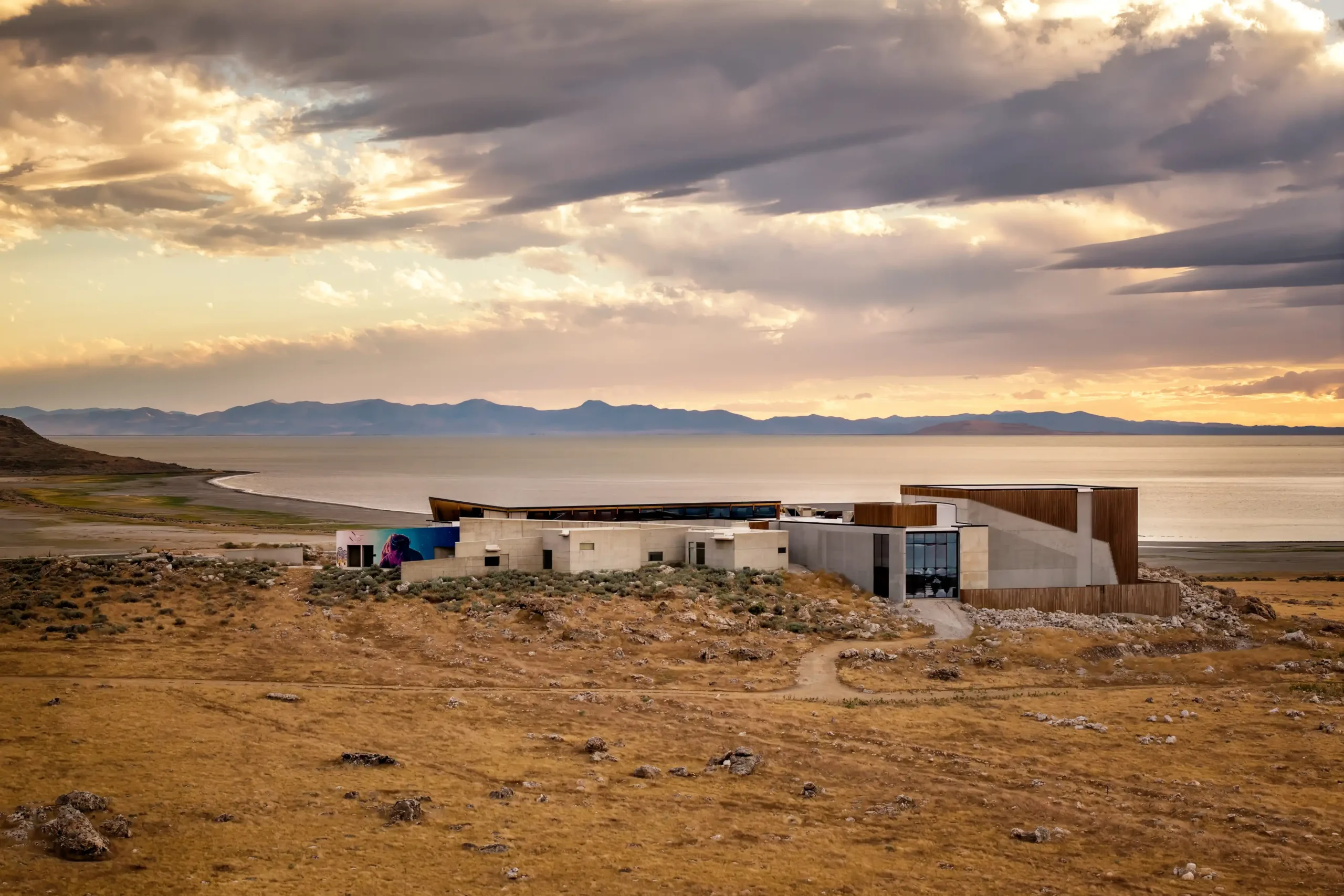 Modern building on dry landscape near a lake, with mountains and dramatic sky at sunset.