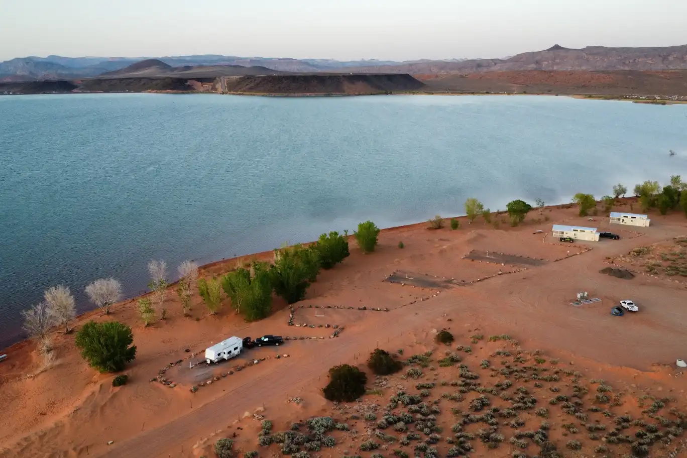 Aerial view of RVs parked by a desert lake with scattered trees and distant hills.