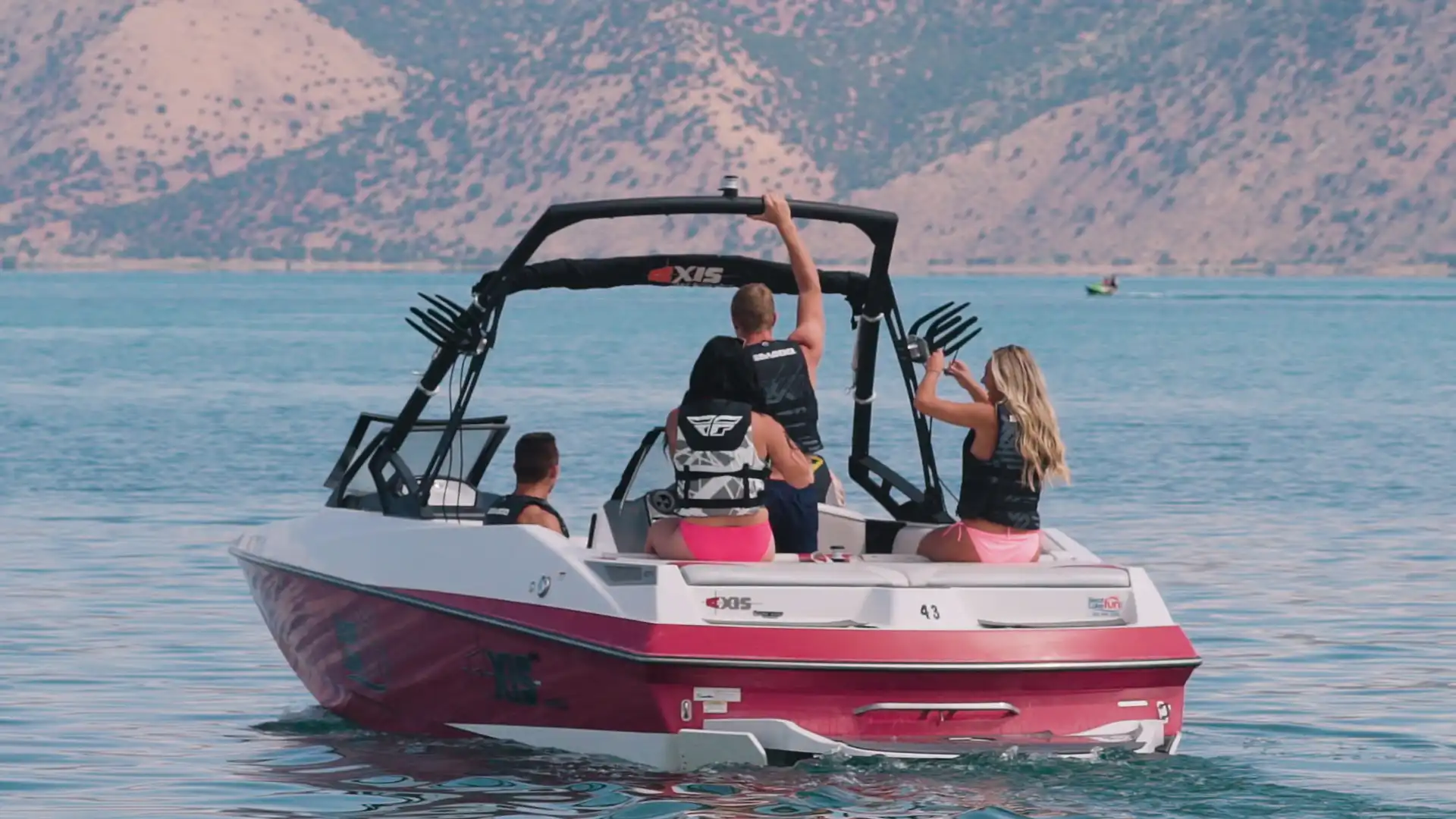 Three people on a red and white speedboat in a lake, with mountains in the background.
