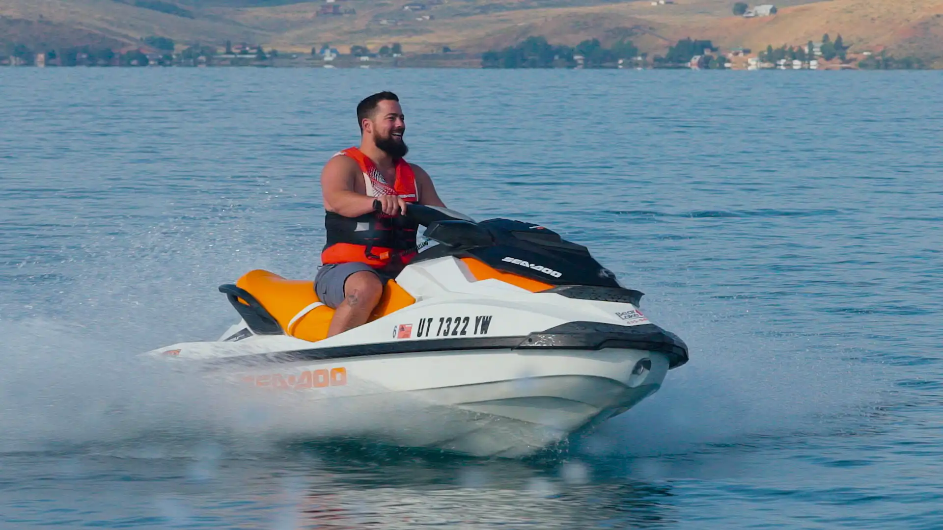 Man in orange life vest rides a white jet ski across a lake with hills in the background.