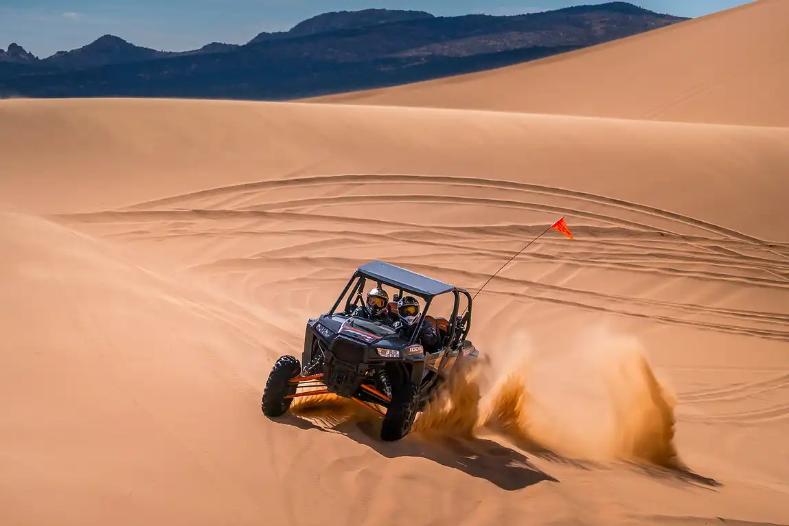 ATV driving through sandy dunes, kicking up dust, with mountains in the background.