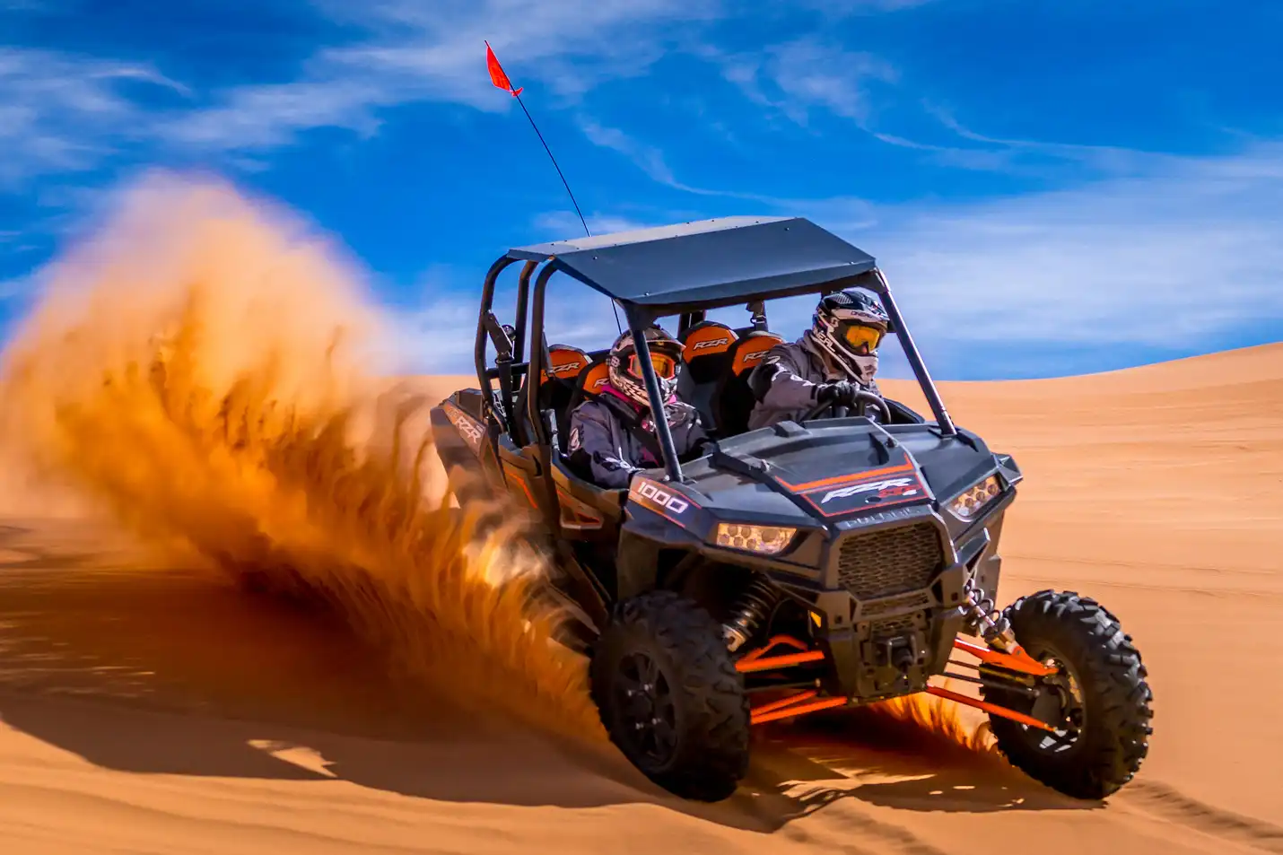ATV speeds through sand dunes, kicking up dust, with two people wearing helmets, blue sky above.
