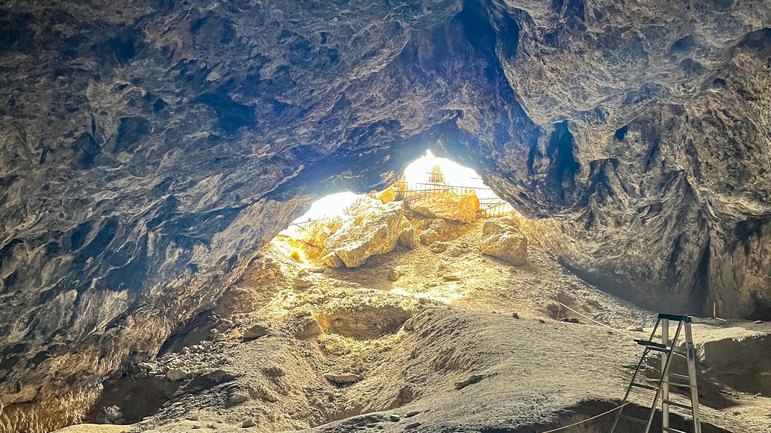 Cave opening with sunlight streaming in, rocks below, and a wooden ladder on the right side.