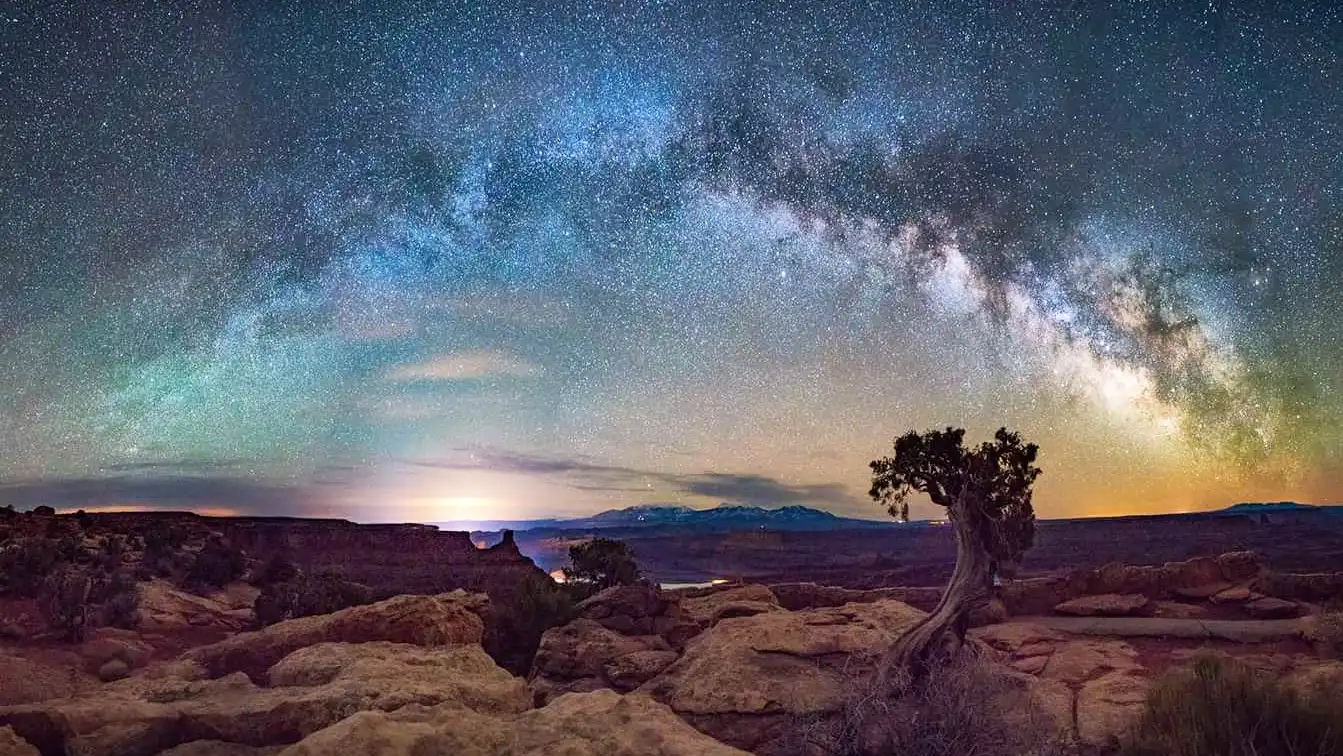 Starry night sky with Milky Way over a rocky desert landscape and a lone tree.