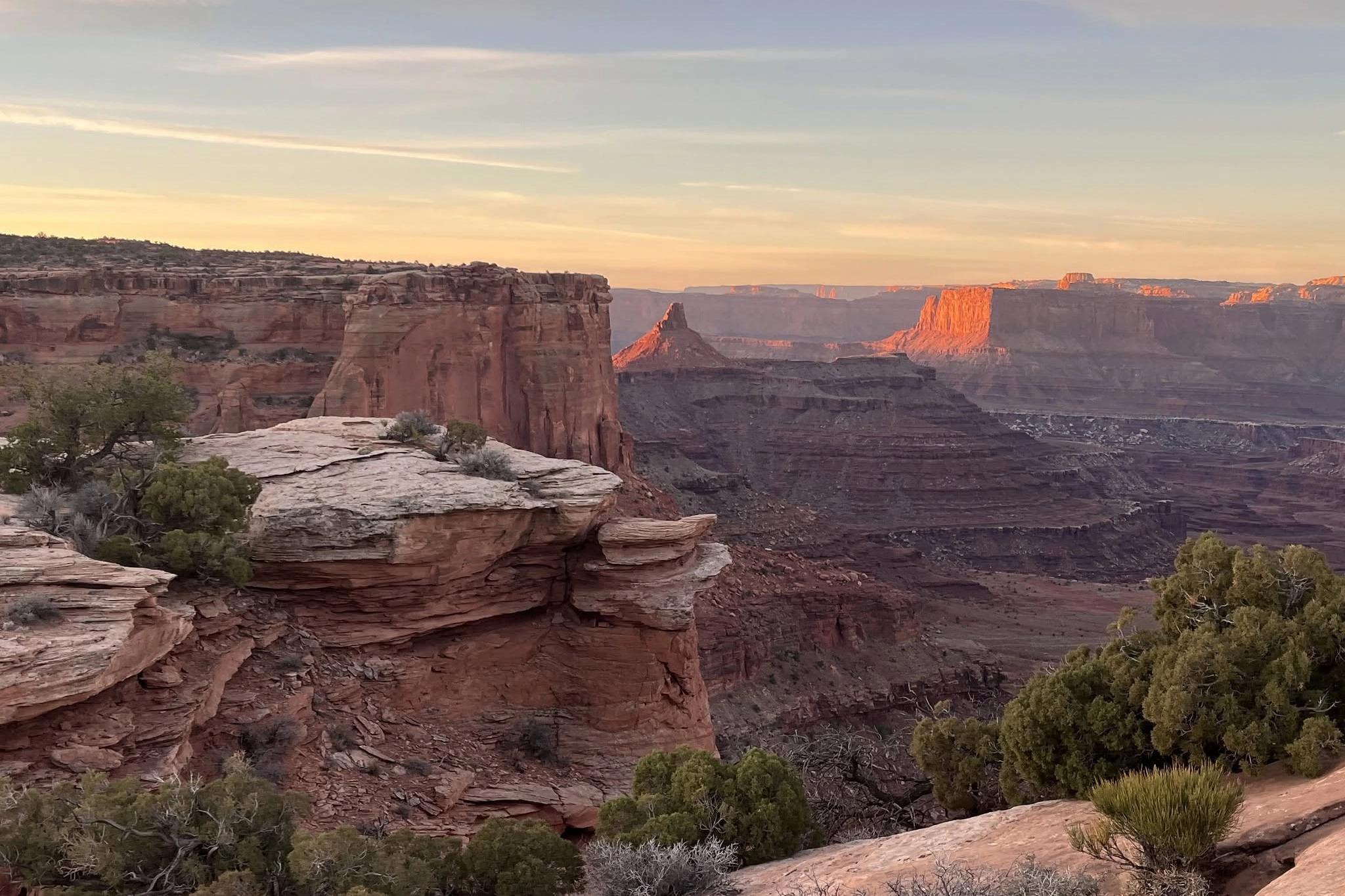 Sunset over red rock canyons with layered cliffs and greenery in the foreground.