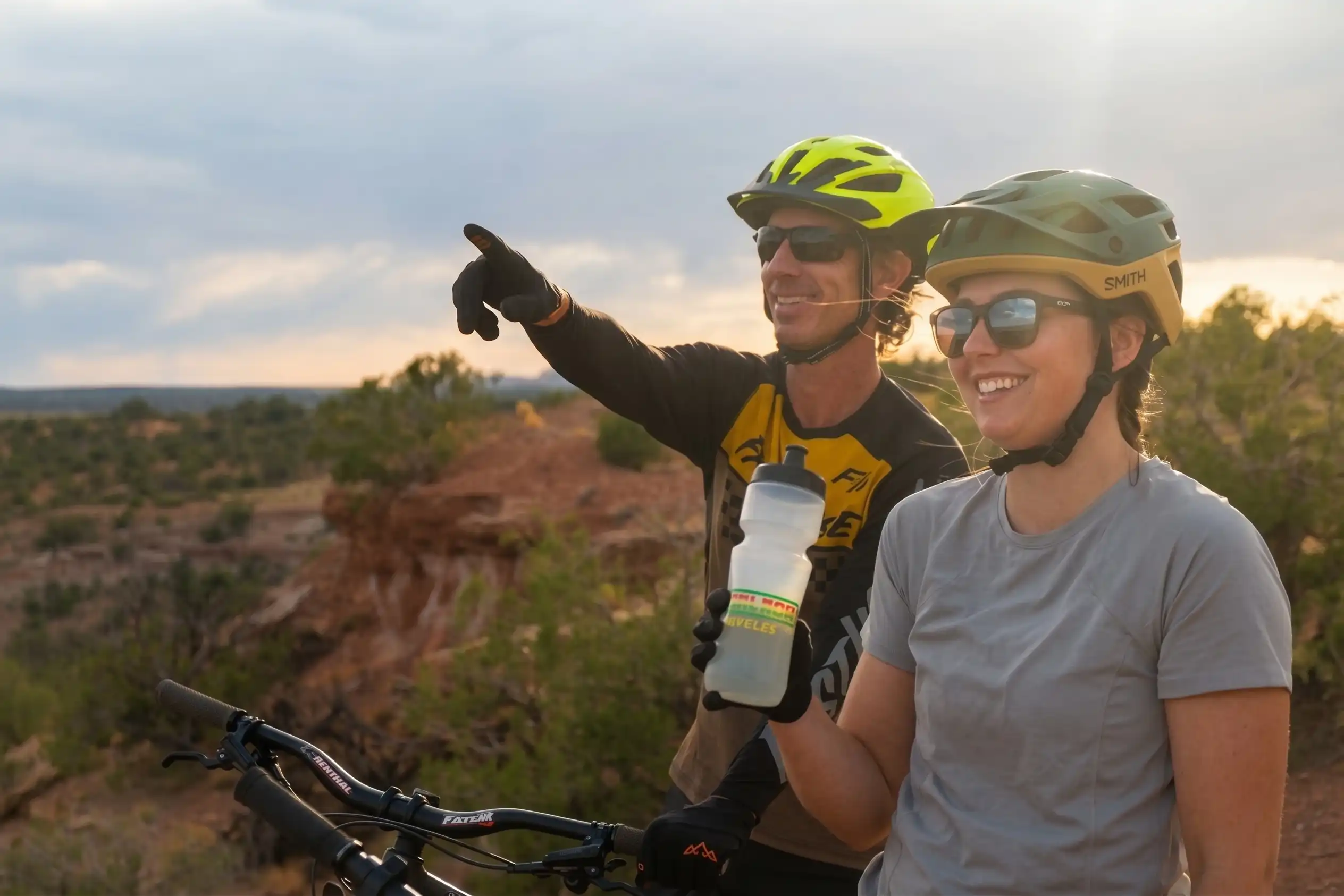 Two cyclists in helmets and sunglasses smile, one points while the other holds a water bottle.
