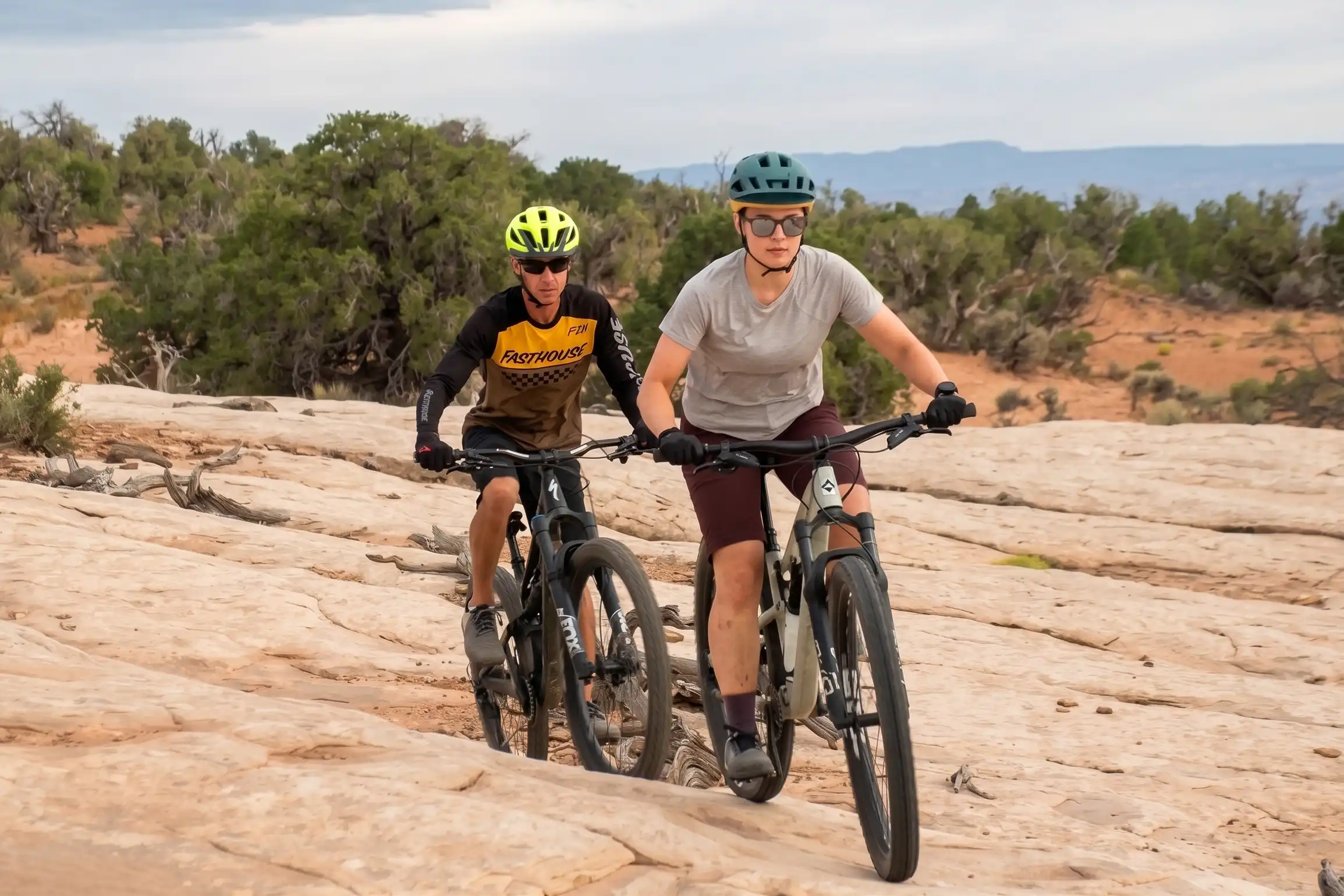 Two people wearing helmets ride mountain bikes on a rocky desert trail with shrubs nearby.