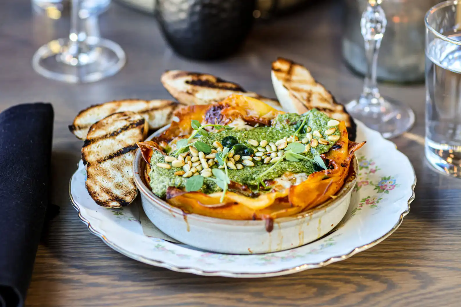 Baked lasagna with pesto, pine nuts, herbs, and toasted bread slices on a floral plate.