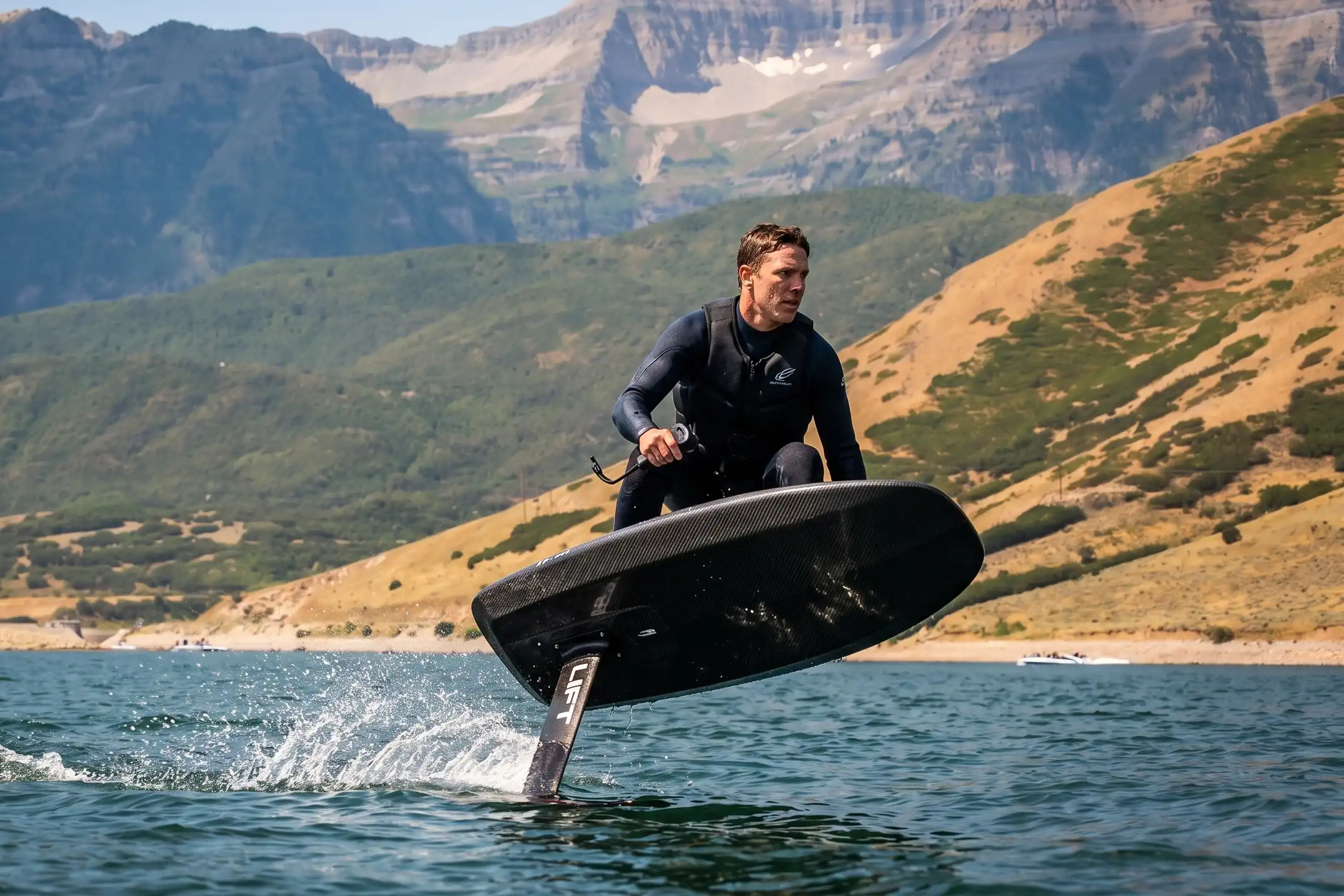 Man rides a hydrofoil surfboard on a lake with mountains in the background.