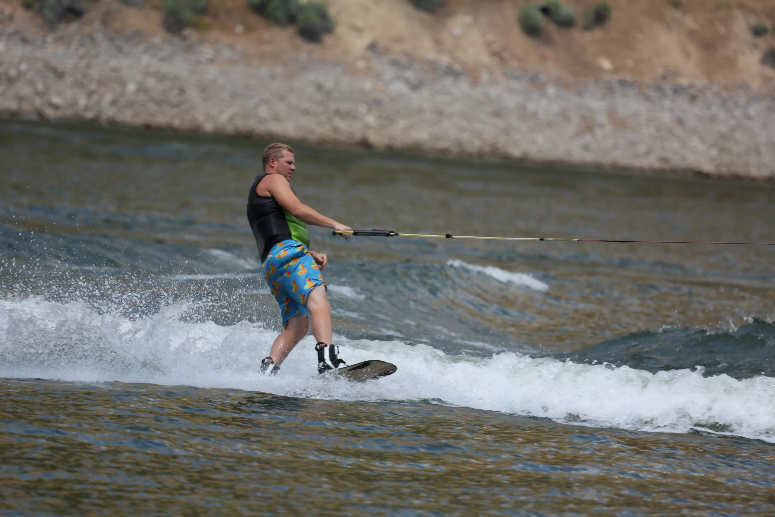 Person wakeboarding on a lake, wearing a black vest and colorful shorts.
