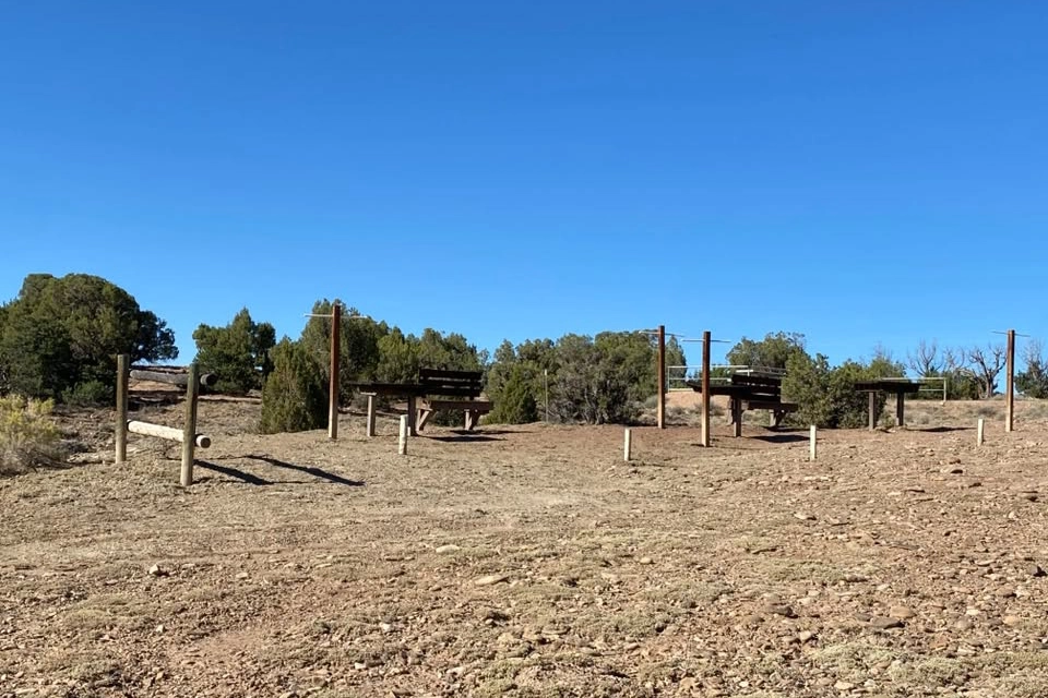 Wooden picnic tables on a dry, rocky hill with scattered shrubs under a clear blue sky.