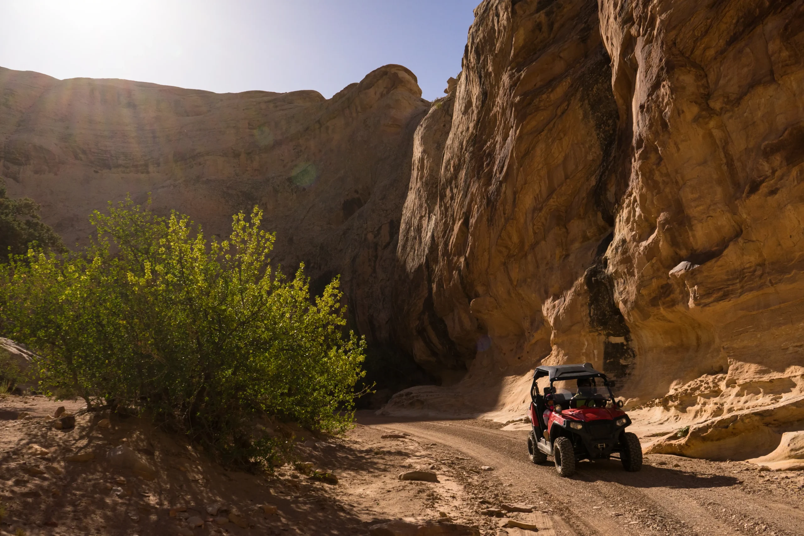 Red ATV drives on a dirt road between tall canyon walls under a bright, sunny sky.