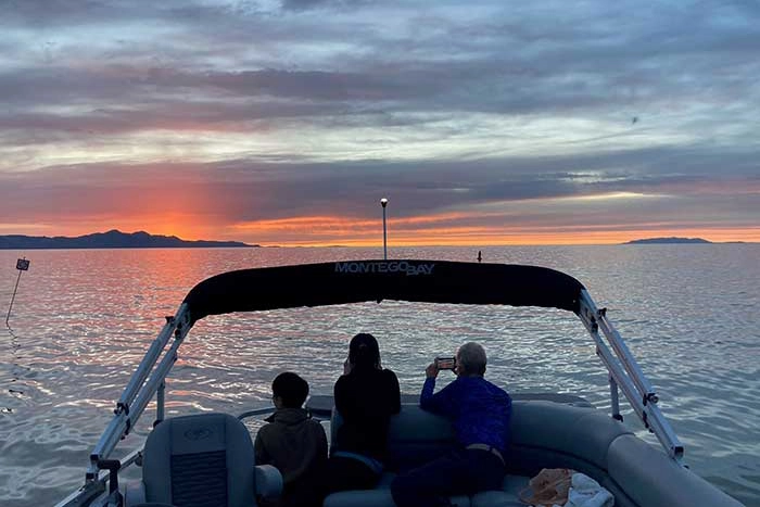 Three people on a boat watch and photograph a vibrant sunset over calm water.