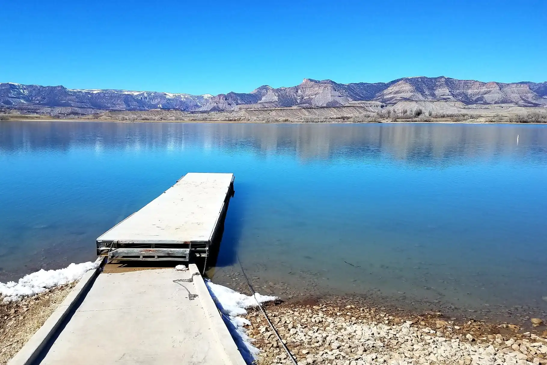 A concrete dock extends into a calm lake with mountains in the background under a clear sky.
