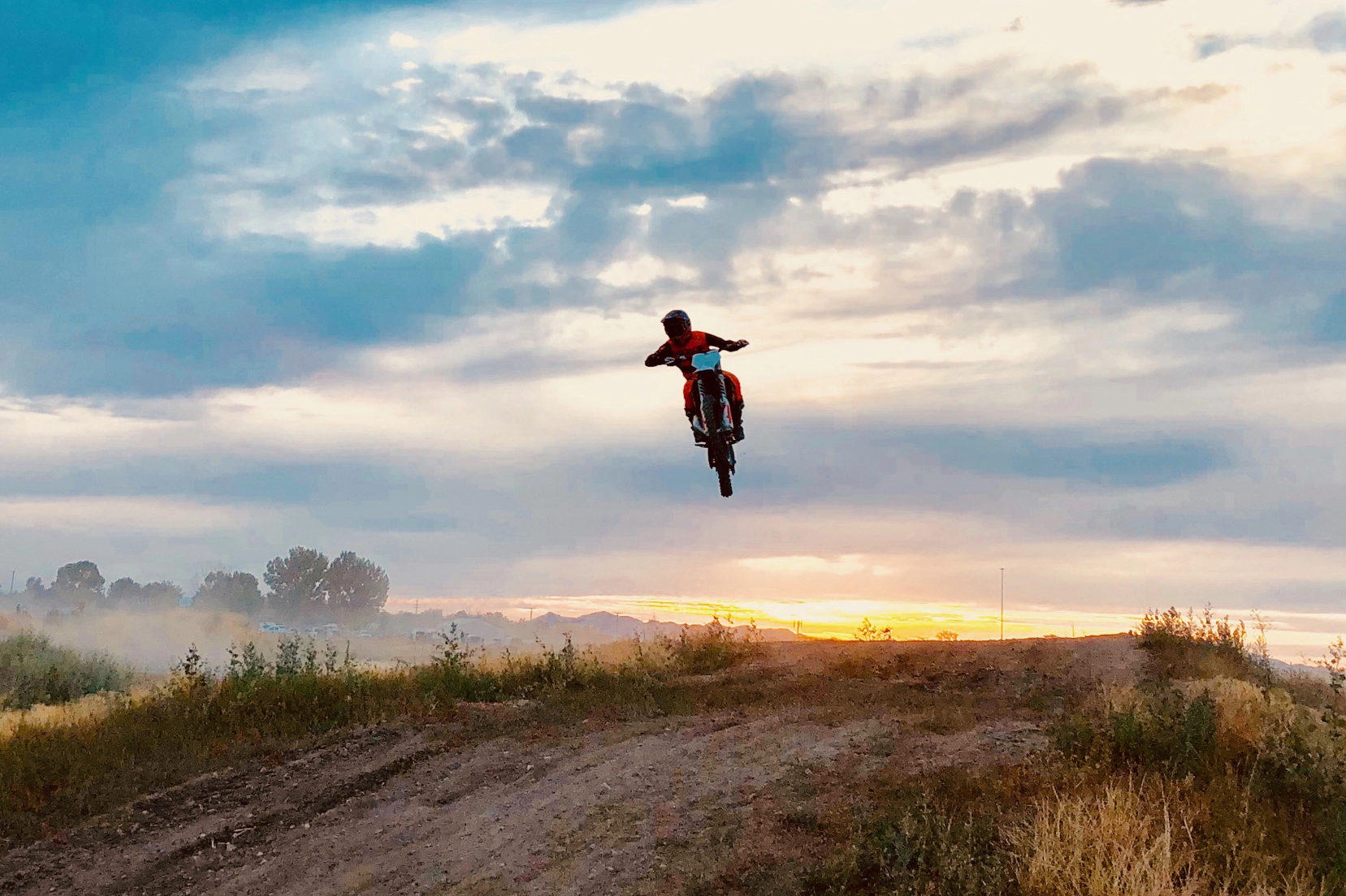 A person on a dirt bike jumps off a hill at sunset under a cloudy sky.