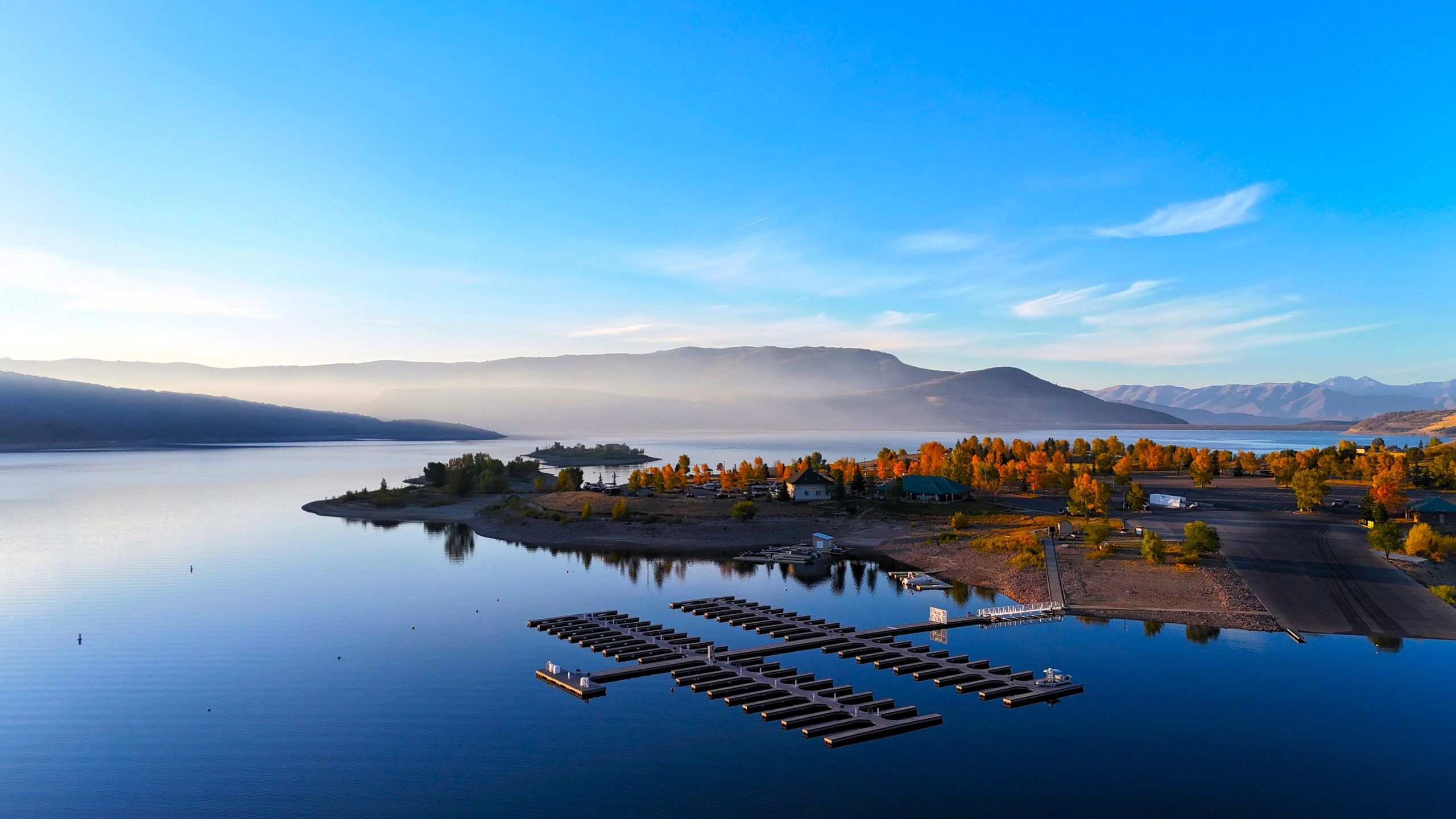 Small marina on a calm lake with fall trees and distant mountains under a blue sky.