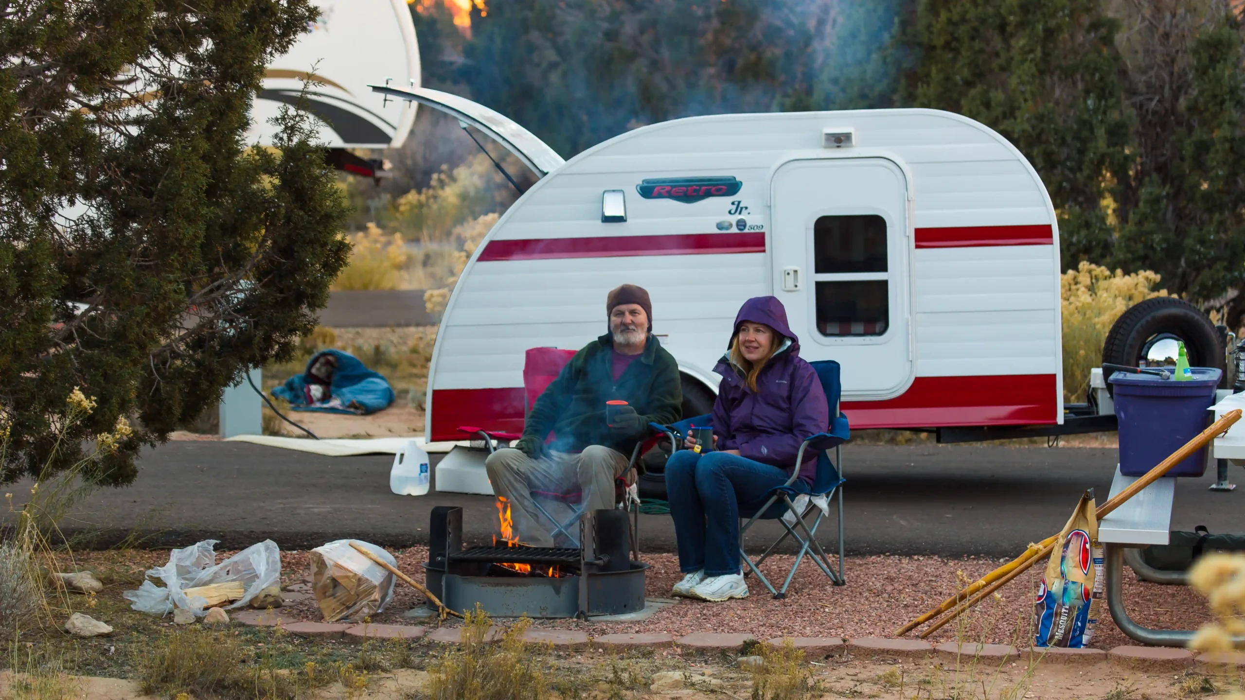 Two people sit by a campfire near a small trailer, smiling and enjoying the outdoors.
