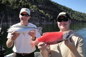 Two men smiling, holding a silver fish and a red fish by a lake with trees and cliffs.