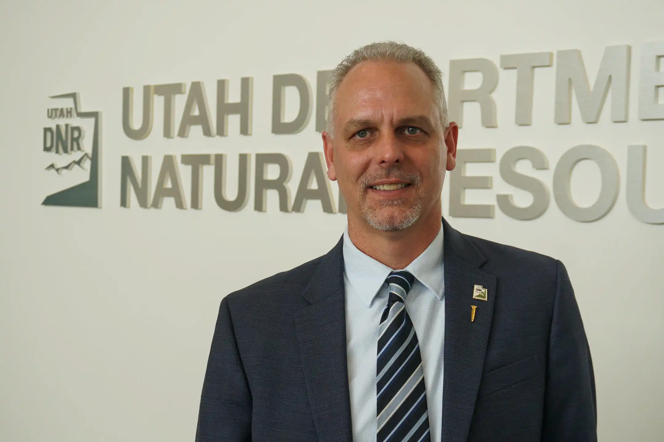 Man in suit standing before Utah Department of Natural Resources sign, smiling at camera.