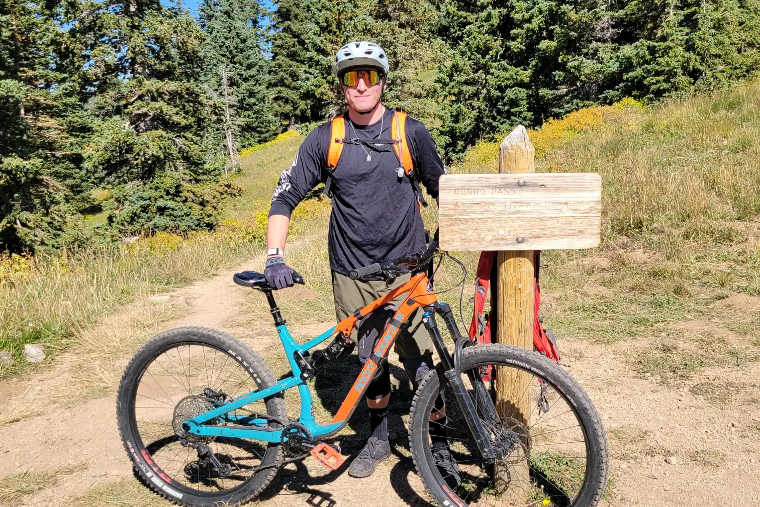 Person in biking gear stands with mountain bike next to a wooden trail sign in a forest.