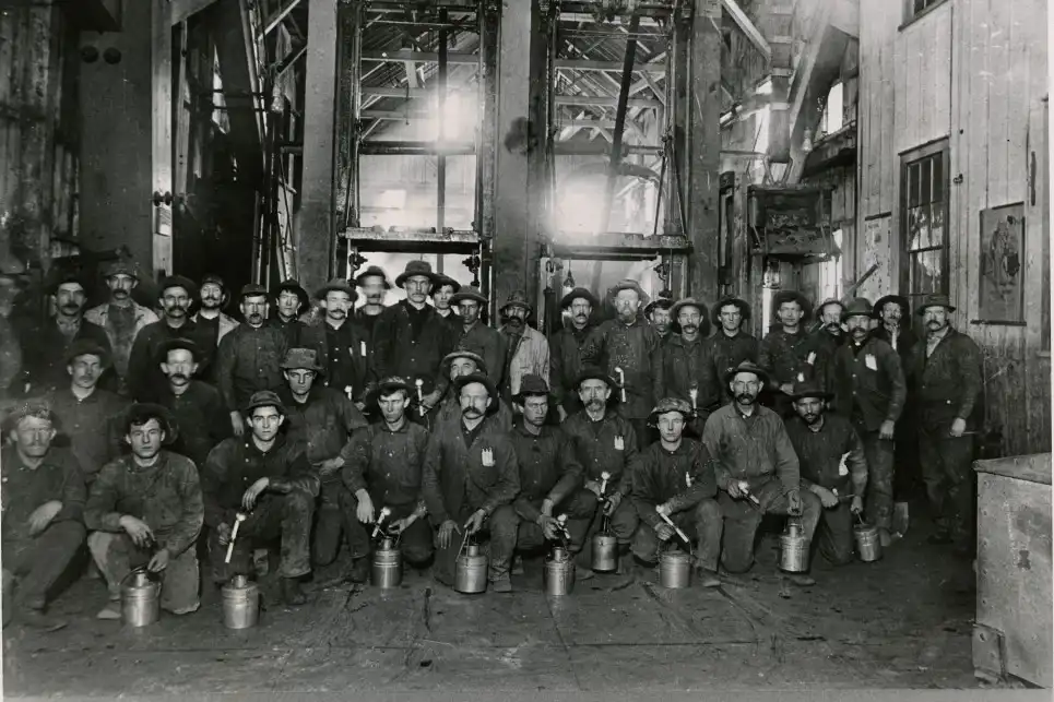 A group of miners pose indoors, holding lunch pails and wearing work clothes and hats.