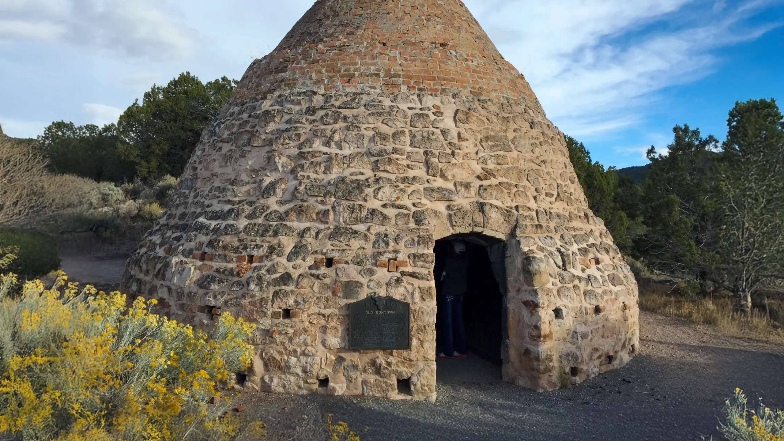Cone-shaped stone structure with a small entrance, surrounded by trees and wildflowers.