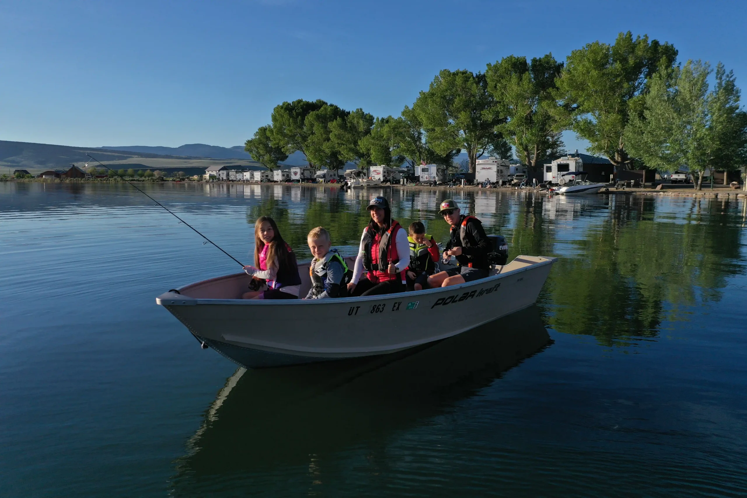 Four people in a small boat on a lake, wearing life jackets, surrounded by trees and RVs.