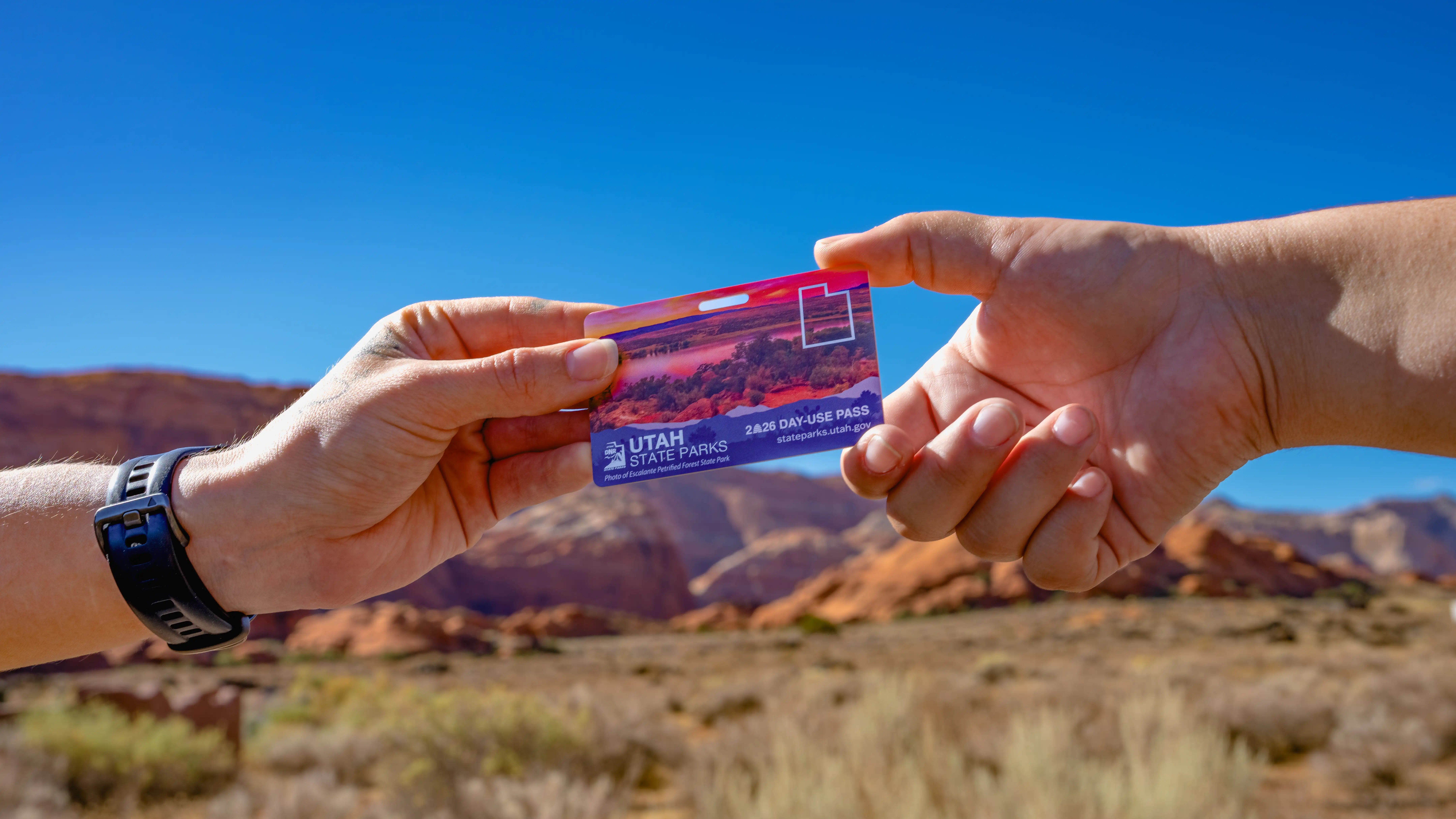 Two hands exchanging a 2026 Utah State Parks day-use pass against a desert landscape.