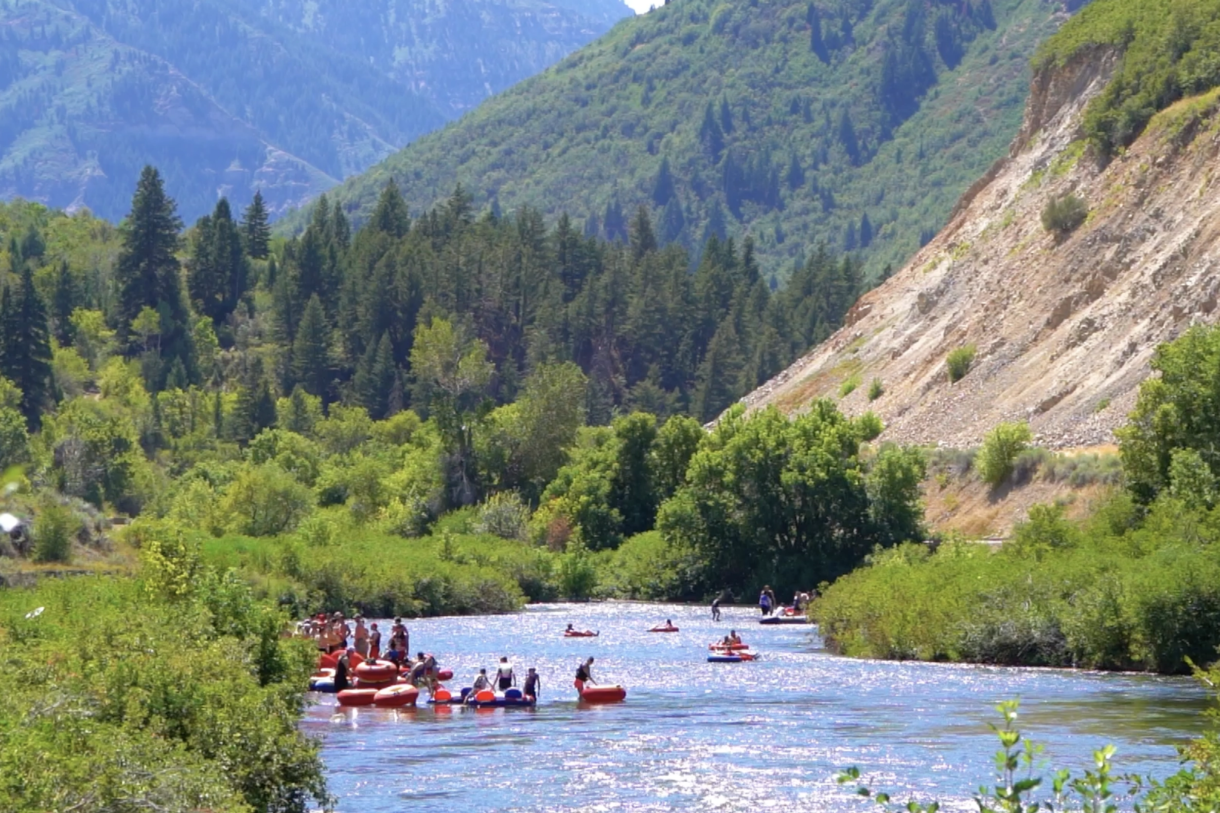 People tubing on a river surrounded by green trees and mountains under a clear sky.