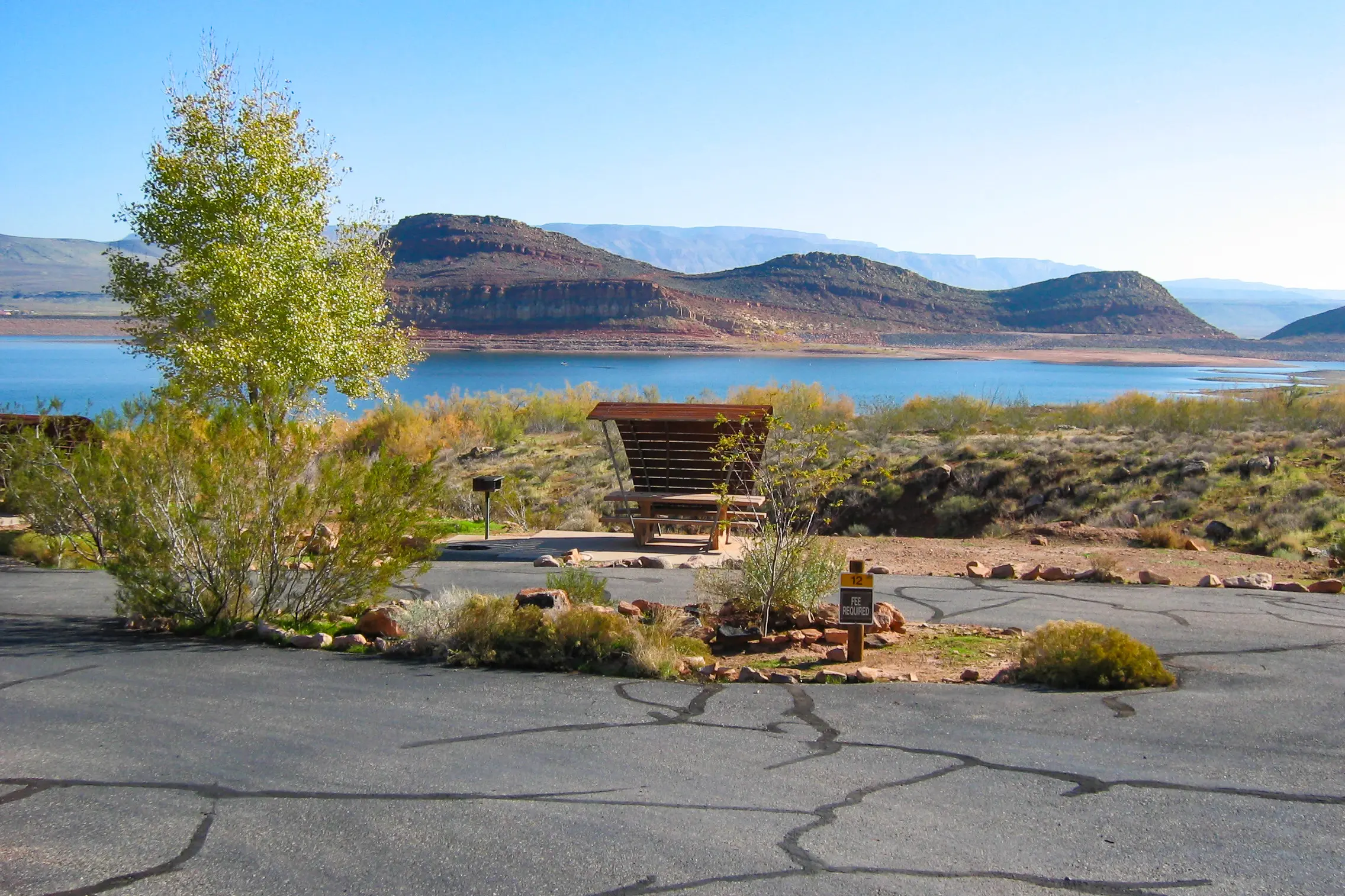 Lake with red rock mesas, picnic area, and tree beside an asphalt path under a clear blue sky.
