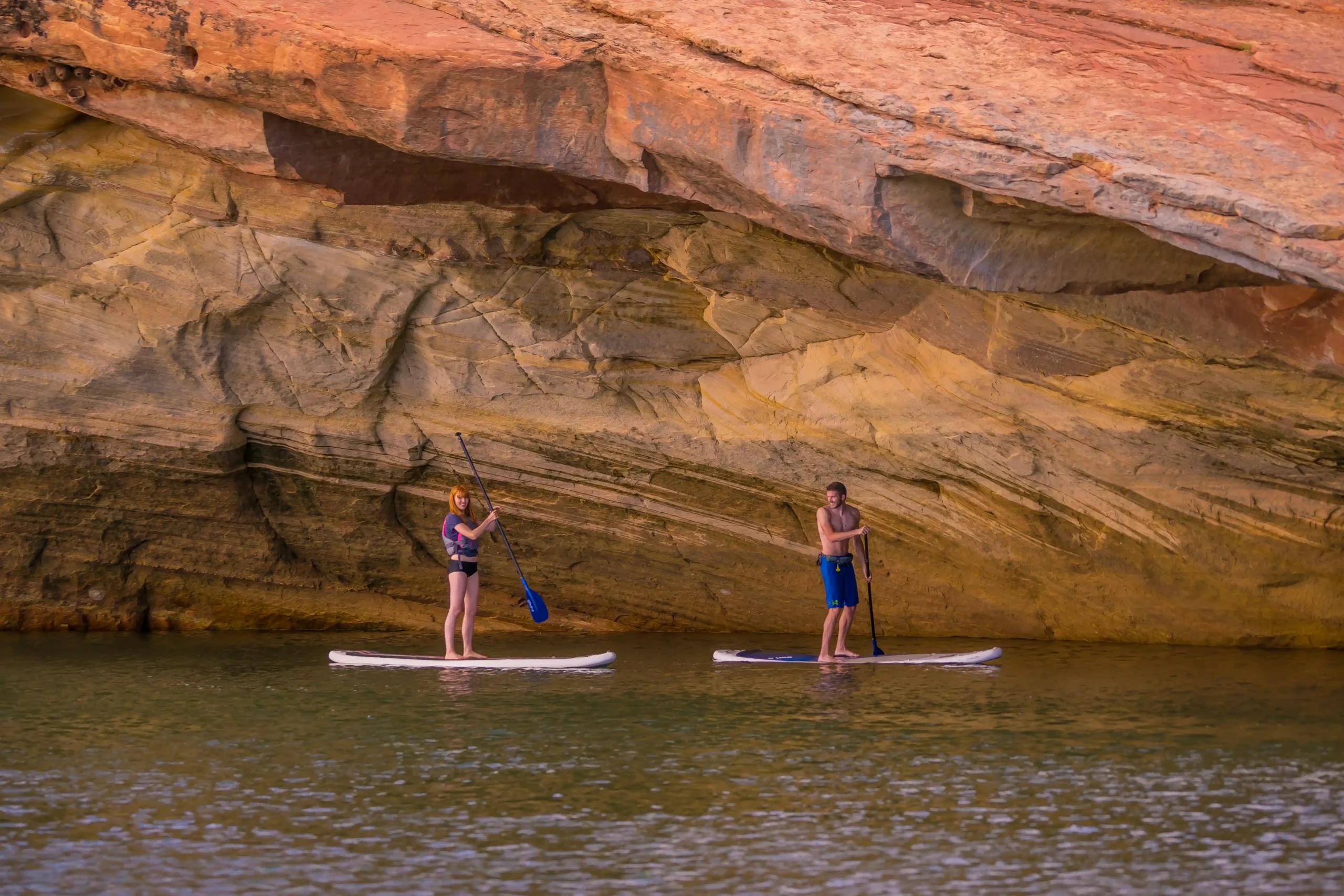 Two people paddleboarding on calm water near a large, colorful rock formation.