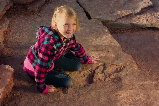 Smiling girl in plaid jacket sits on rocks near a large dinosaur footprint.
