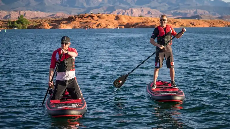 Two men paddleboarding on a lake, wearing life vests, with rocky hills in the background.