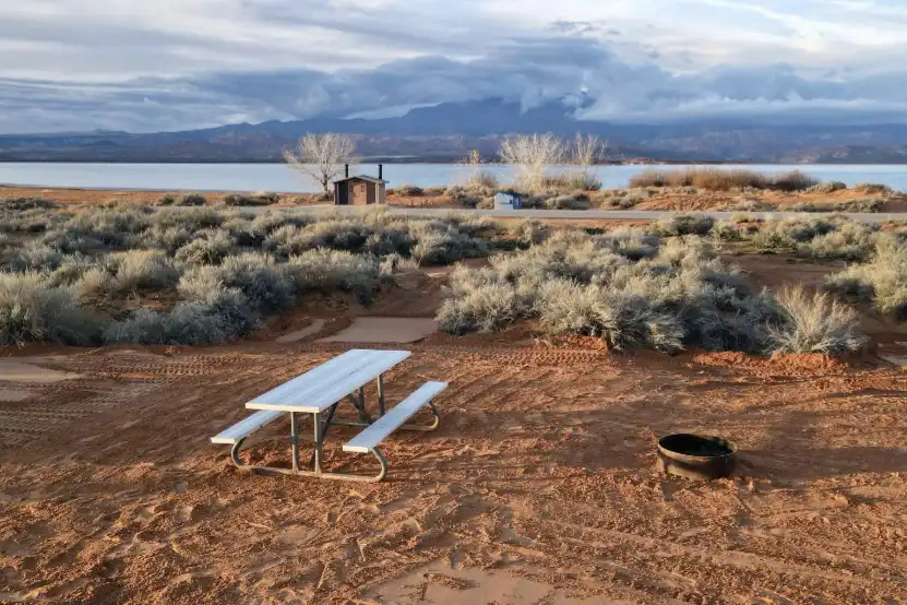 White picnic table and fire ring on sandy ground near a lake with shrubs and mountains behind.