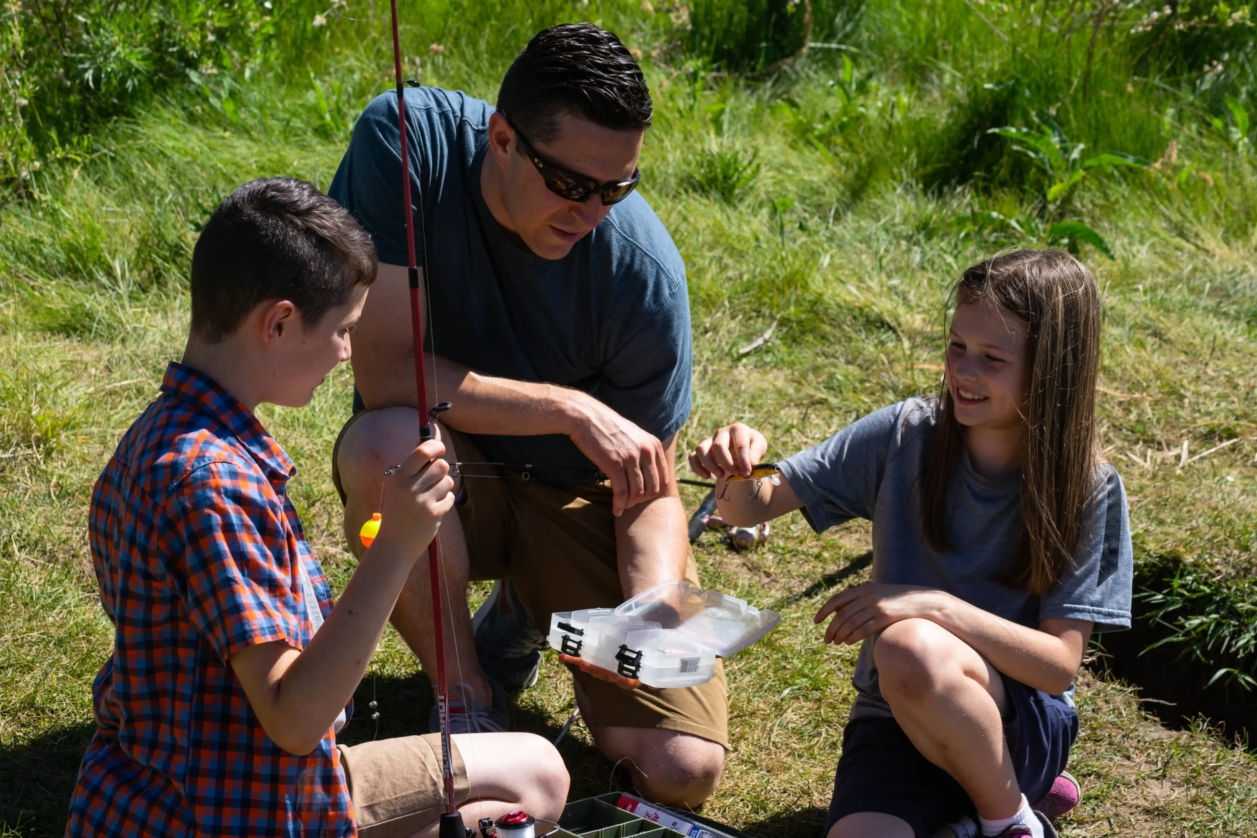 Two kids and an adult fishing, preparing tackle in the grass on a sunny day.