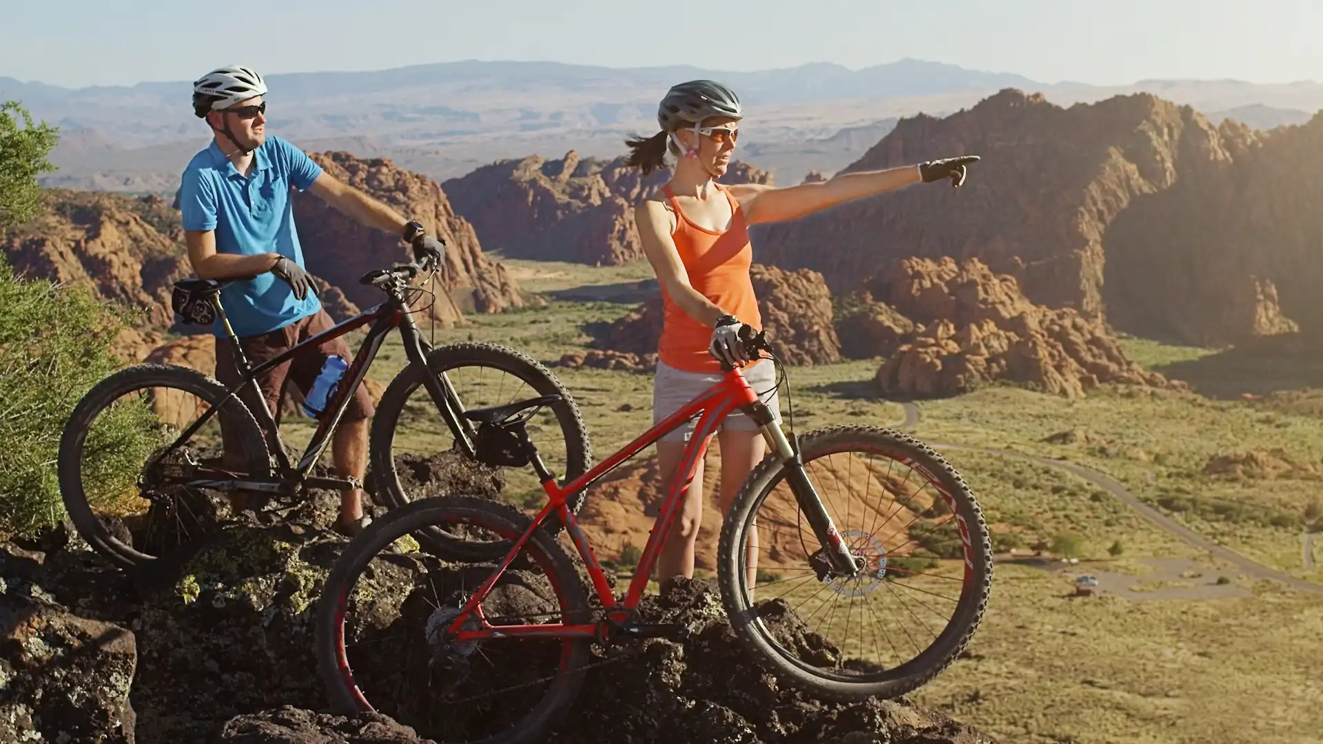 Two cyclists in helmets on rocky terrain, with one pointing toward distant mountains.