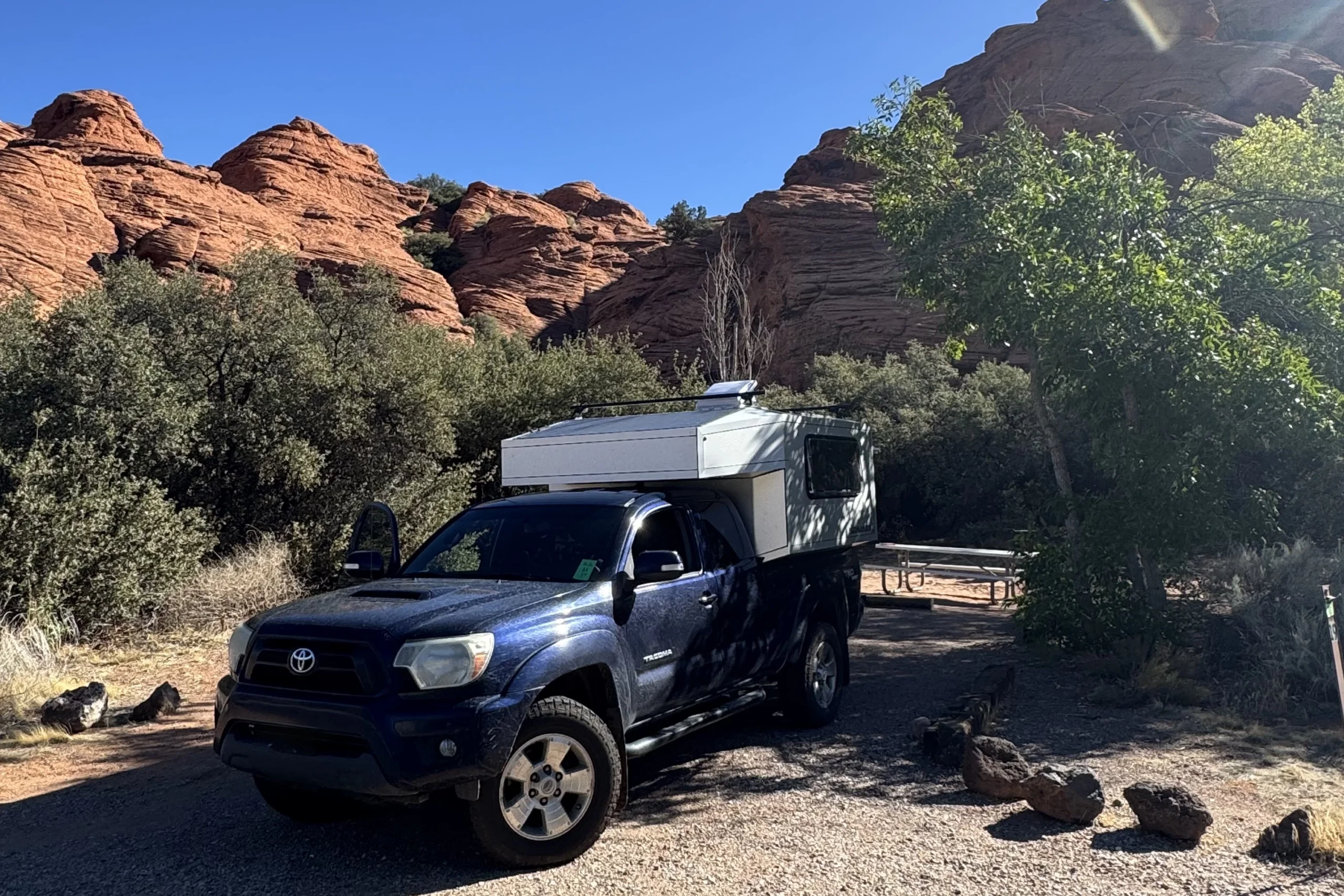 A dark blue truck with a camper is parked at a campsite near red rock formations and trees.