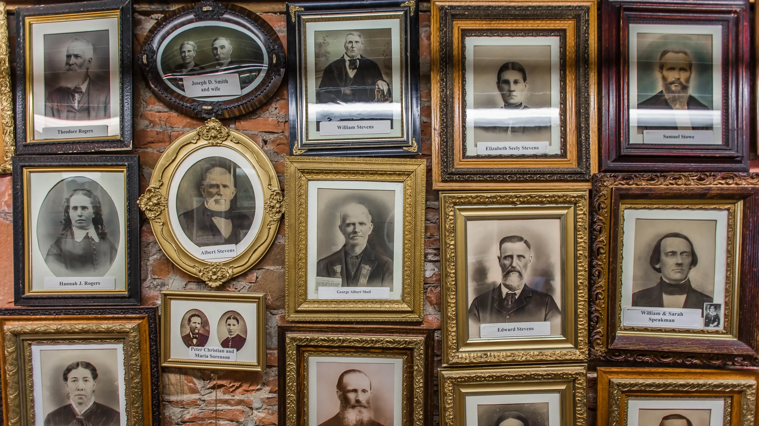 Framed old-fashioned portraits arranged closely on a brick wall, featuring men and women.