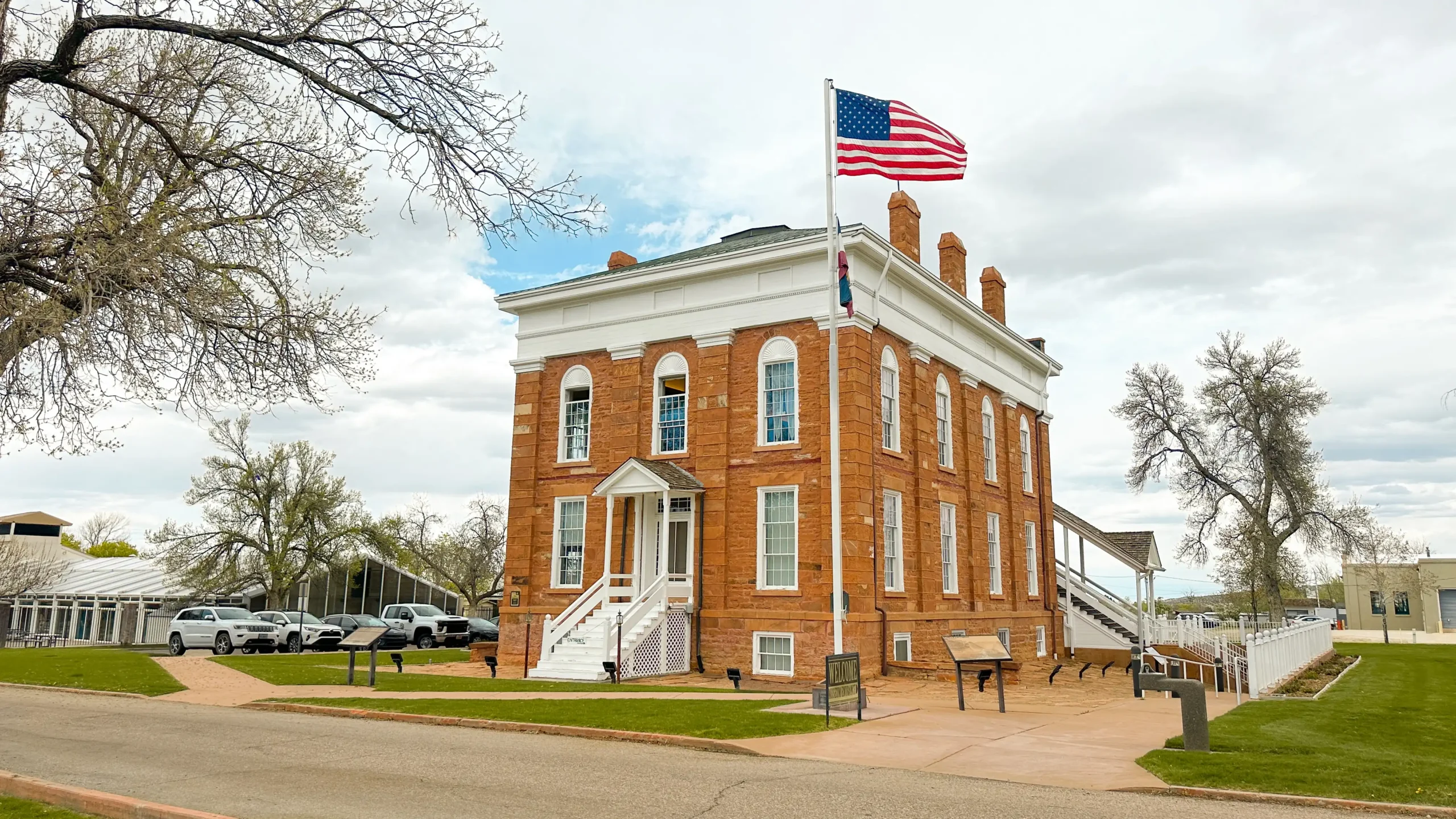 Red brick courthouse with US flag, white trim, stairs, and cars parked nearby on a cloudy day.