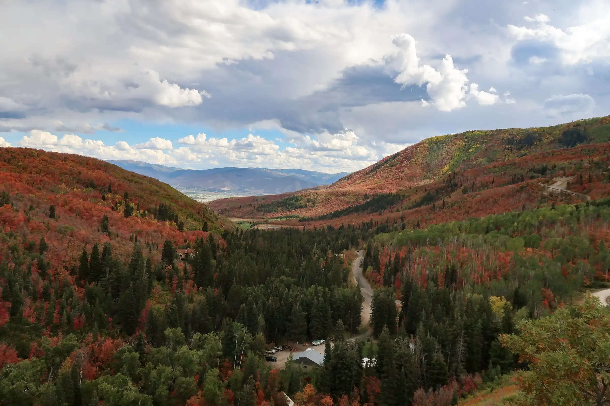 Valley with green and red trees, winding road, hills, and a partly cloudy sky.