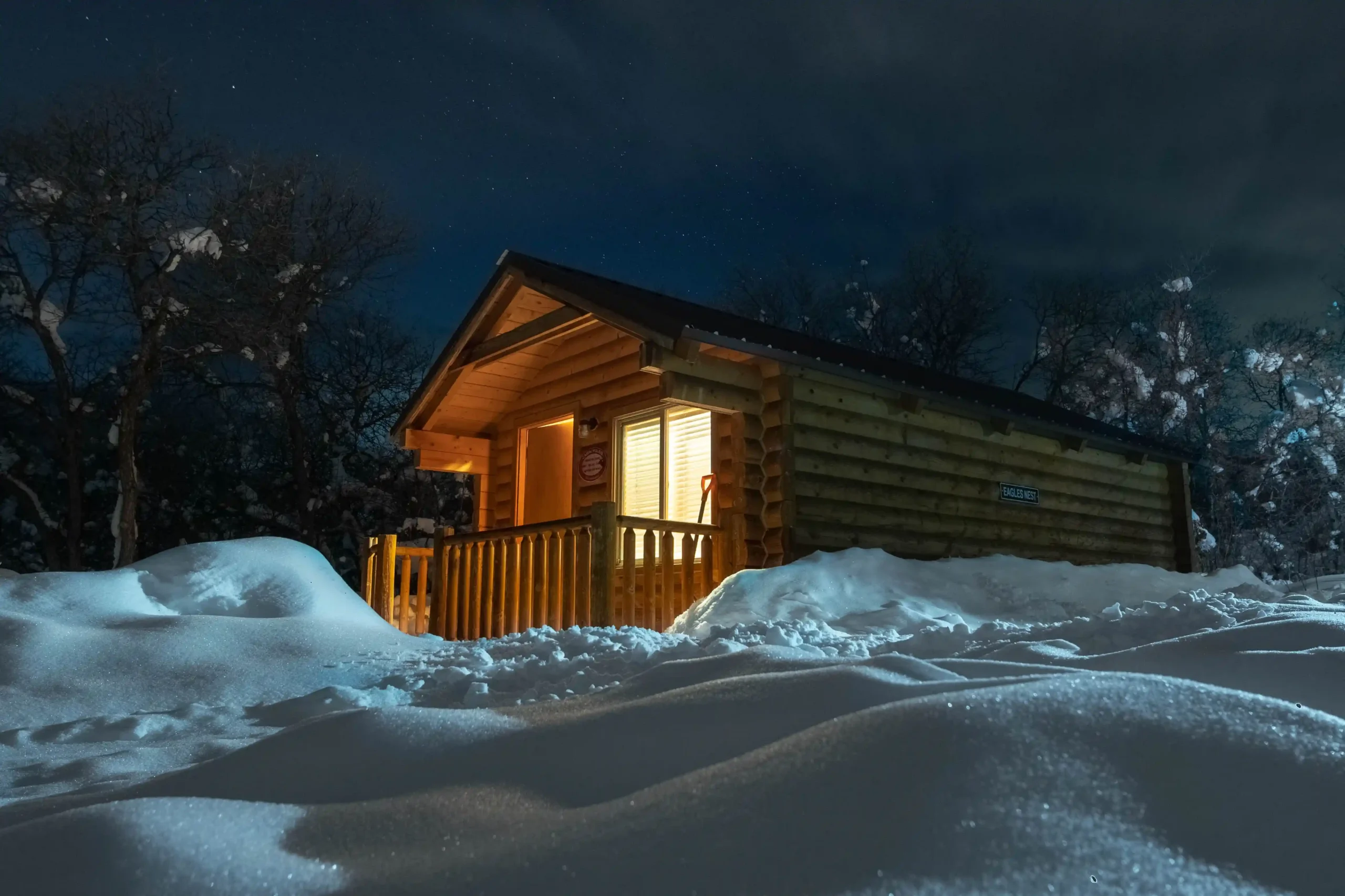 A snowy cabin glows warmly at night, surrounded by trees under a dark sky.