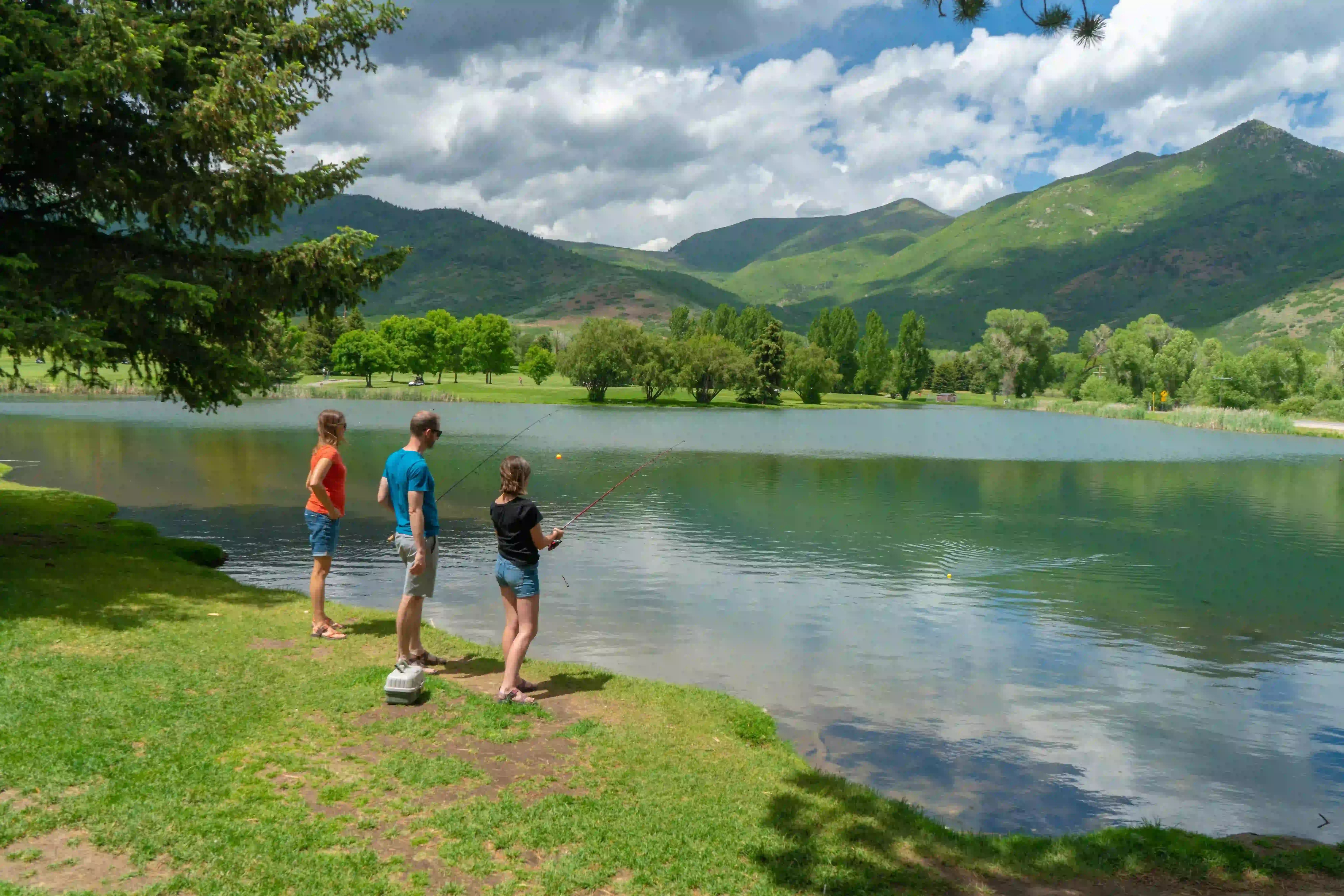 Three people fishing by a scenic lake with mountains and greenery in the background.