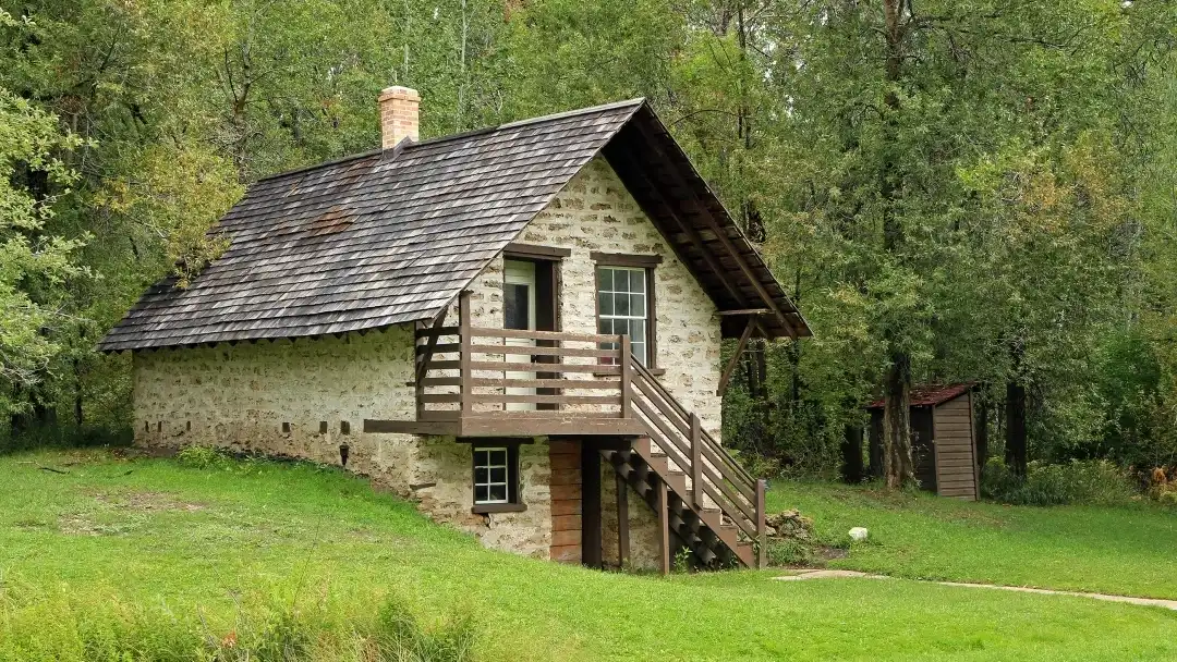 Stone cabin with a wooden porch and stairs, surrounded by green grass and trees.