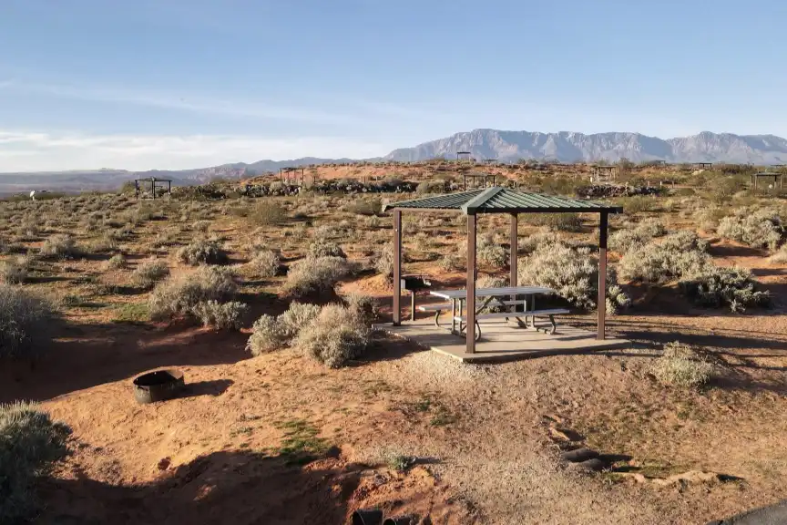 Picnic shelter with benches in a desert landscape, mountains in the background.