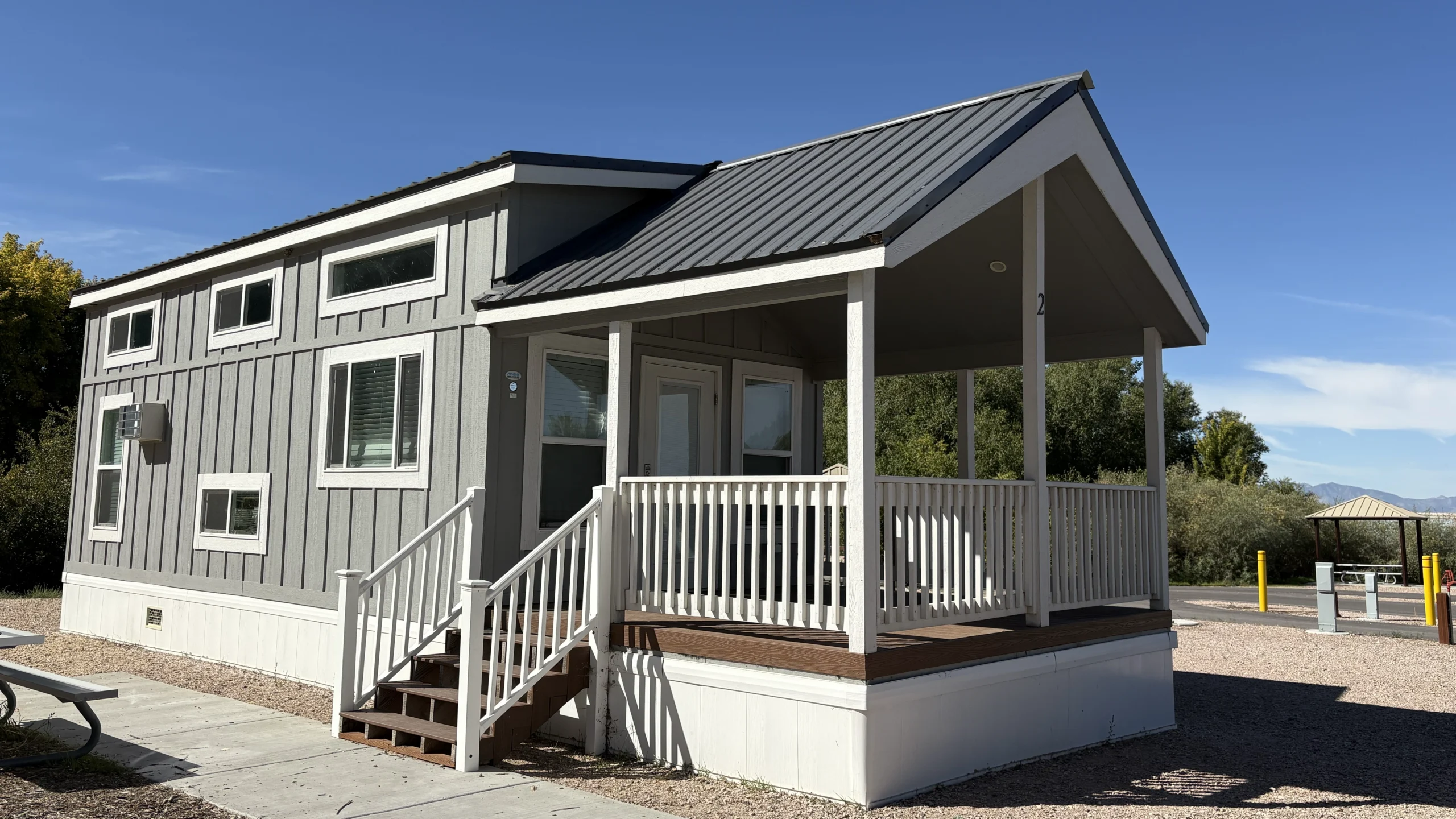 Gray tiny house with a front porch, steps, and railing on a sunny day.