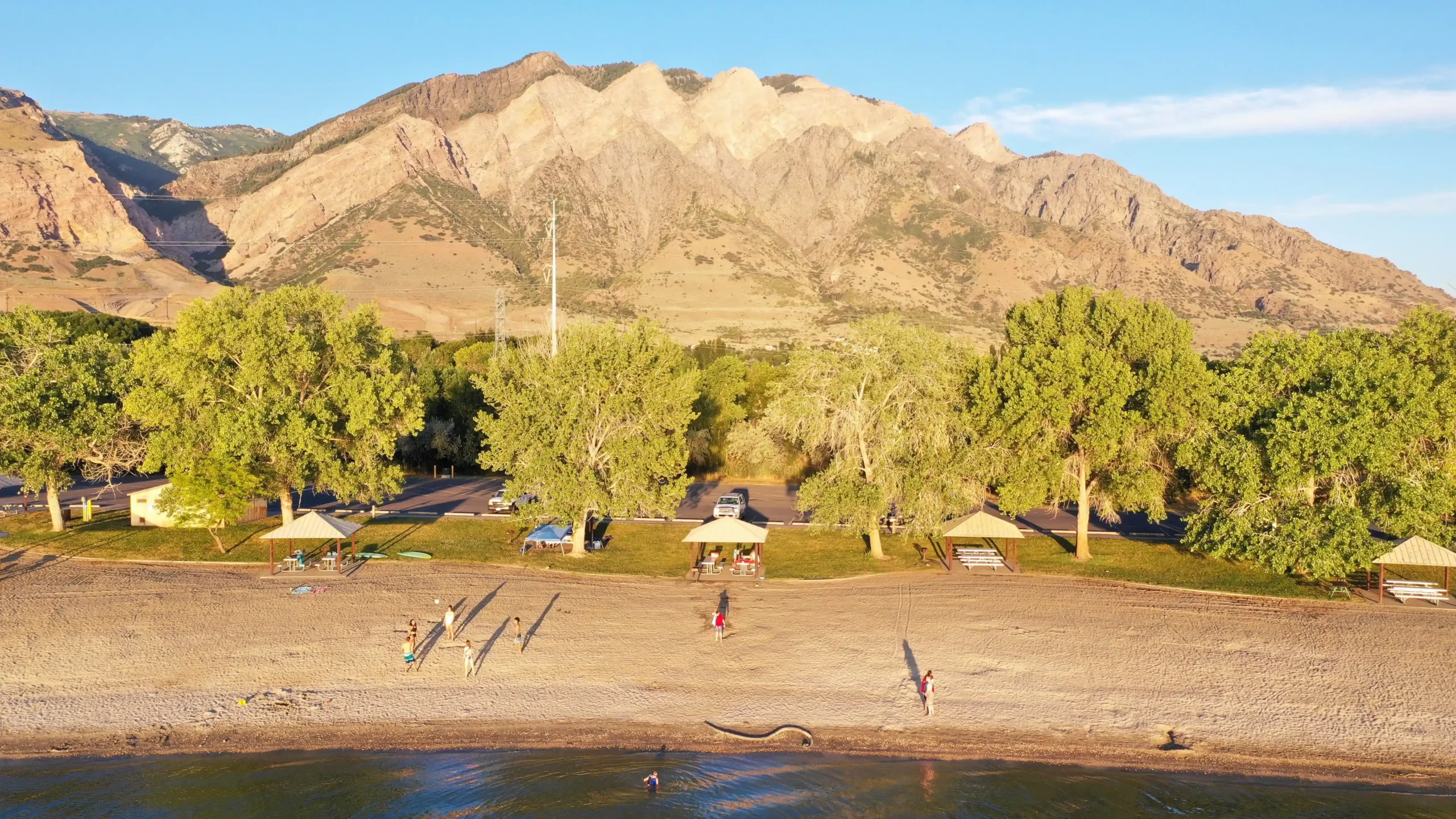 Beach park with trees, pavilions, people, and a mountain in the background under clear skies.