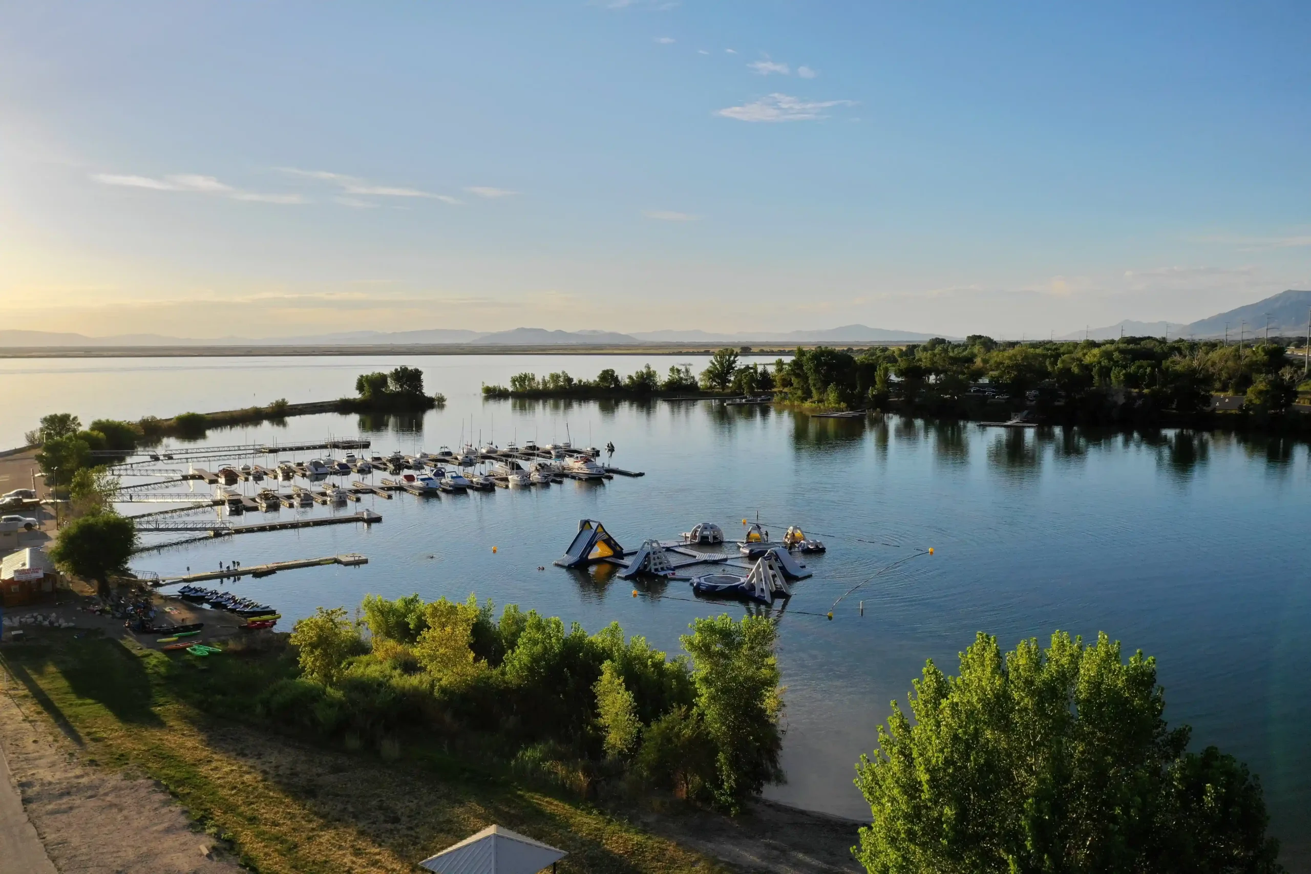 A marina with boats and floating inflatables on a calm lake, surrounded by trees and hills.